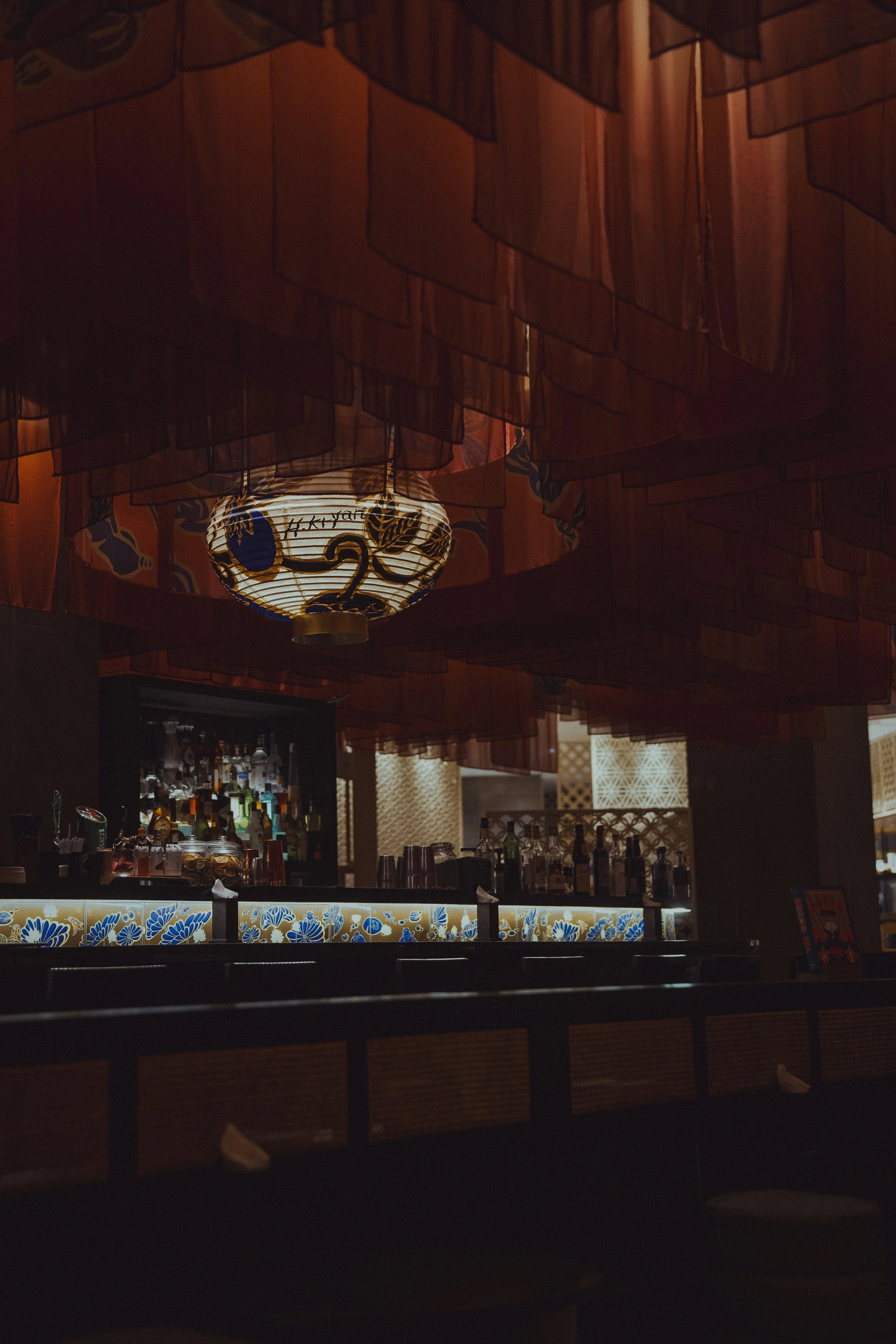 The image shows a dimly lit bar area with bottles of liquor on display, colorful glassware, and a decorative illuminated panel with floral patterns. Overhead, there are layered red and orange fabric curtains and a traditional Japanese-style paper lantern providing lighting.