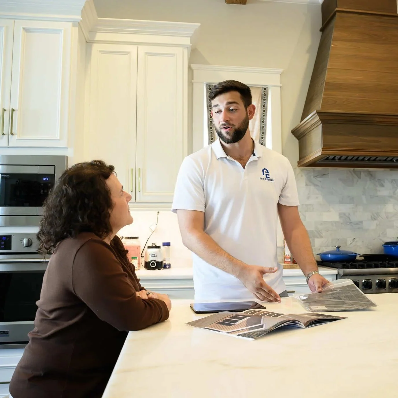 A roofing professional discusses insurance claim documentation with a homeowner at a kitchen counter, reviewing material samples and paperwork together.