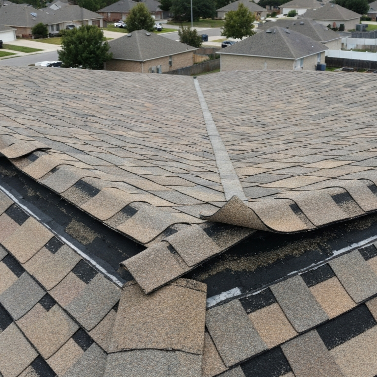 Close-up aerial view of wind-damaged asphalt shingles and lifted ridge cap components on residential roof after severe storm conditions.
