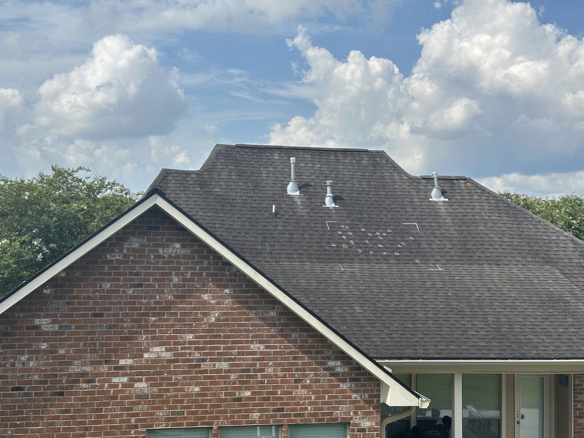 A house with a brick wall and a shingled roof. The roof has three metal vents and a patch of shingles that are missing or damaged.