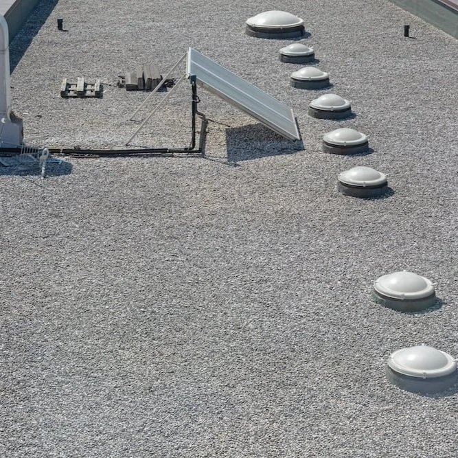Aerial view of a flat gravel roof featuring dome skylights, HVAC units, and a solar panel, highlighting underlayment inspection areas.