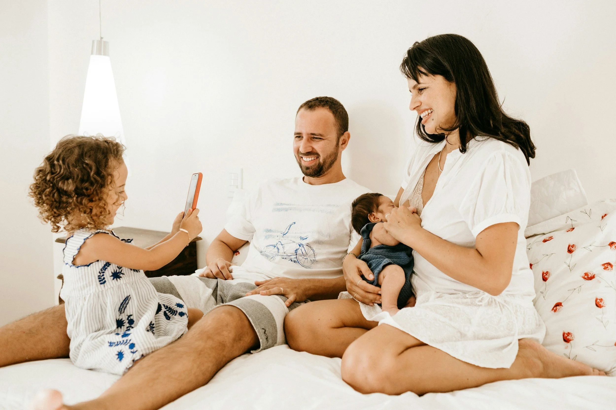 A happy family with a newborn and toddler sitting on a bed together, illustrating what families gain from the Sleepy Mums REST Method.