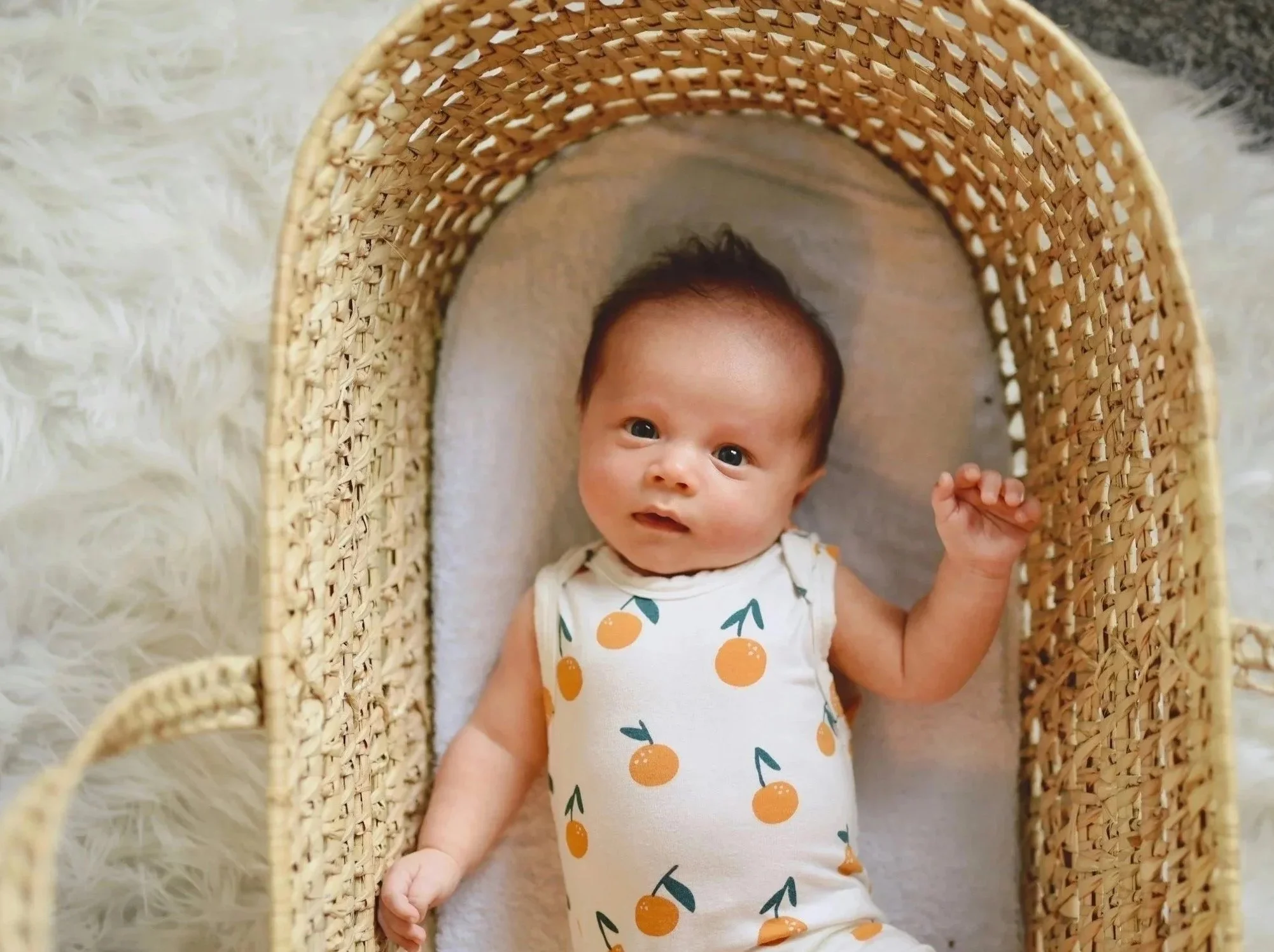 A newborn baby wearing an orange-patterned onesie lying peacefully in a woven Moses basket on a fluffy white rug.