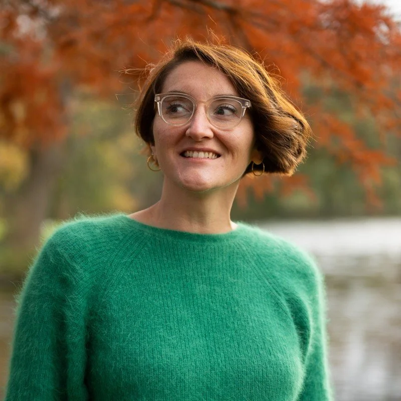 A woman with short brown hair and glasses wearing a green sweater, outdoors in front of autumn trees and a body of water.