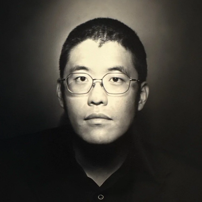Black and white portrait of a young man wearing glasses, with short hair, against a plain background