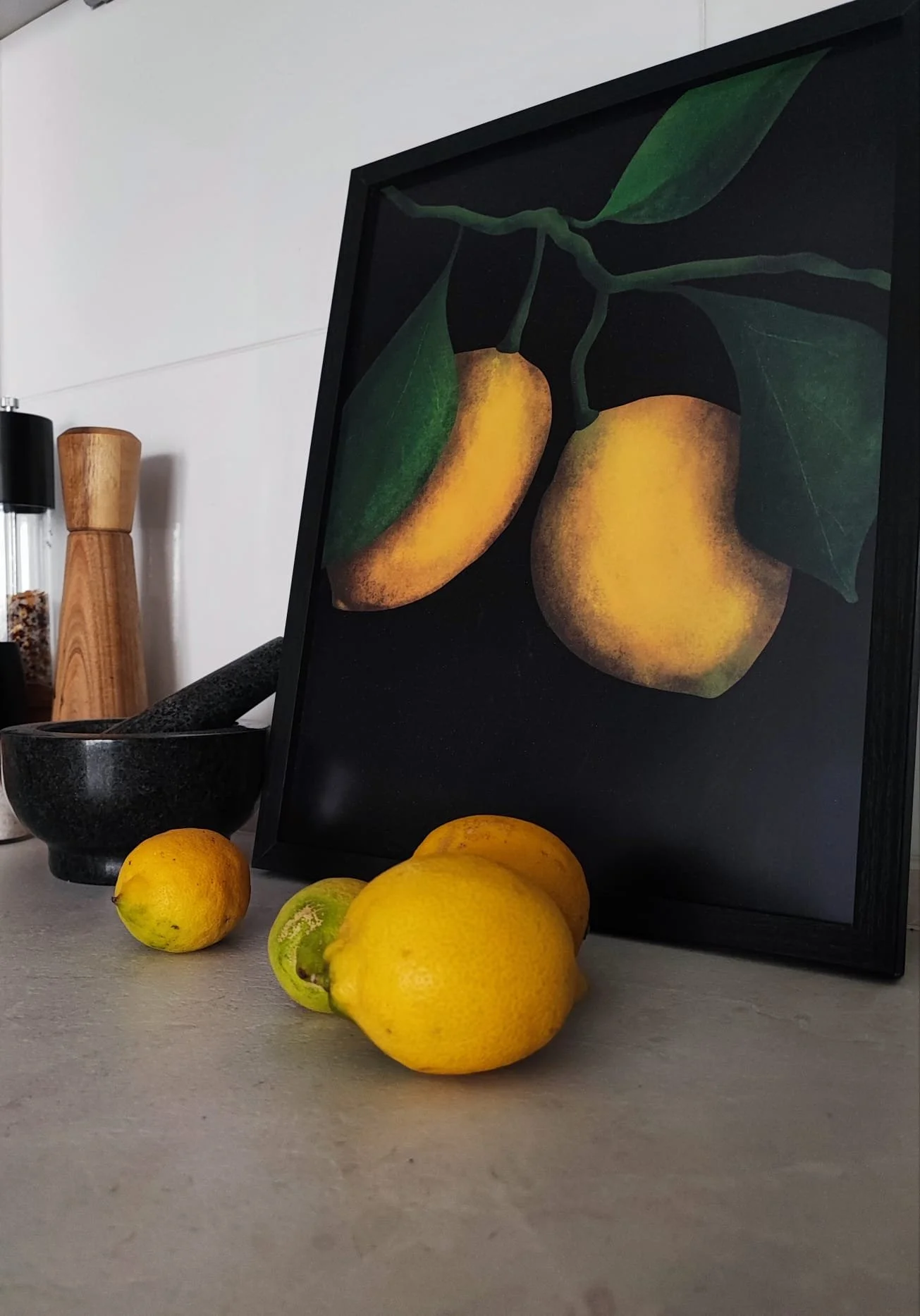 Lemons and limes on a kitchen counter in front of a framed painting of lemons.