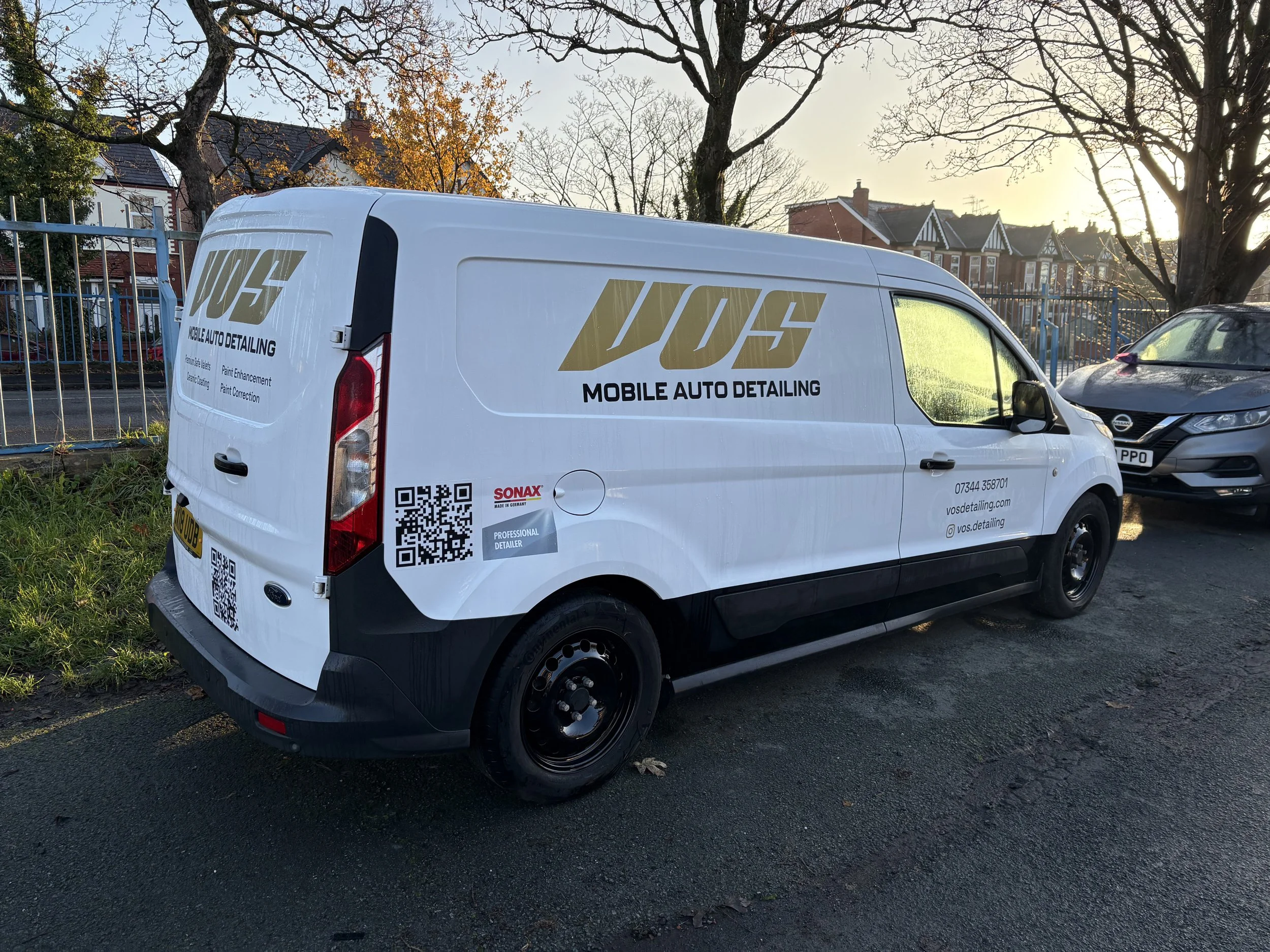 White van with gold and black logo and text advertising mobile auto detailing, parked on a street near a parking lot with trees and residential houses in the background.