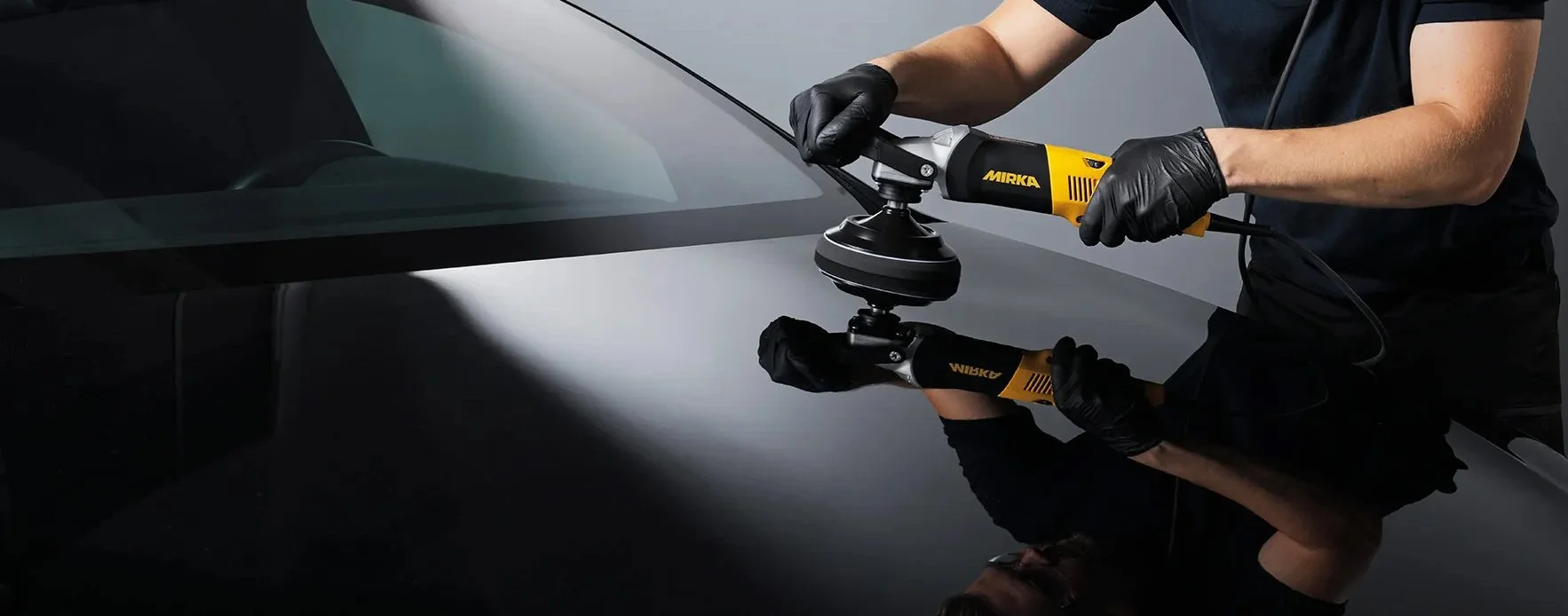 Person polishing a black car with a yellow Mirka power buffer, wearing black gloves, in a dark setting.