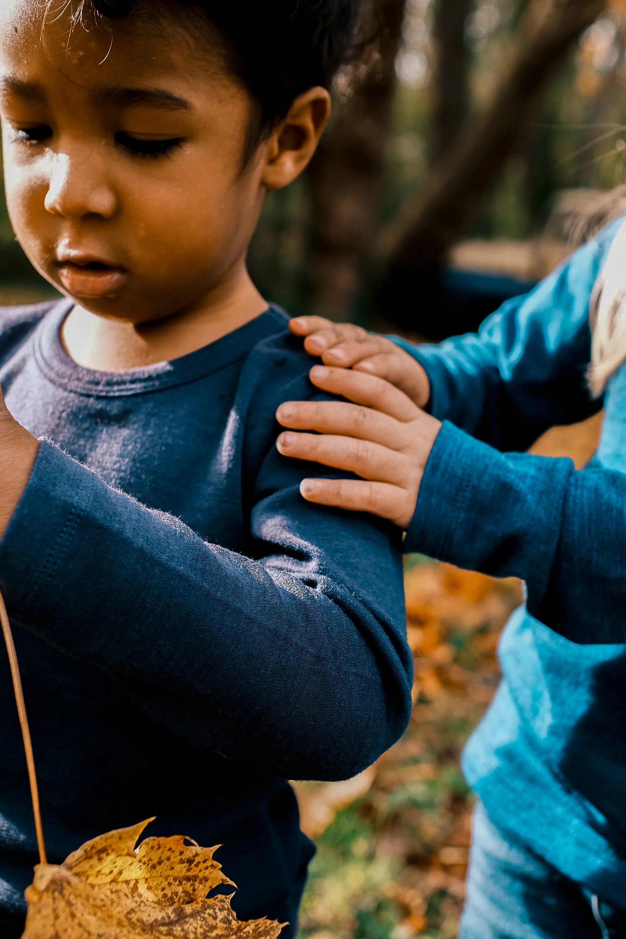 A young boy with dark hair and tan skin wearing a dark blue long-sleeve shirt is being comforted or consoled by another child who is placing a hand on his shoulder. Product photography fashion photography for childrens brands.