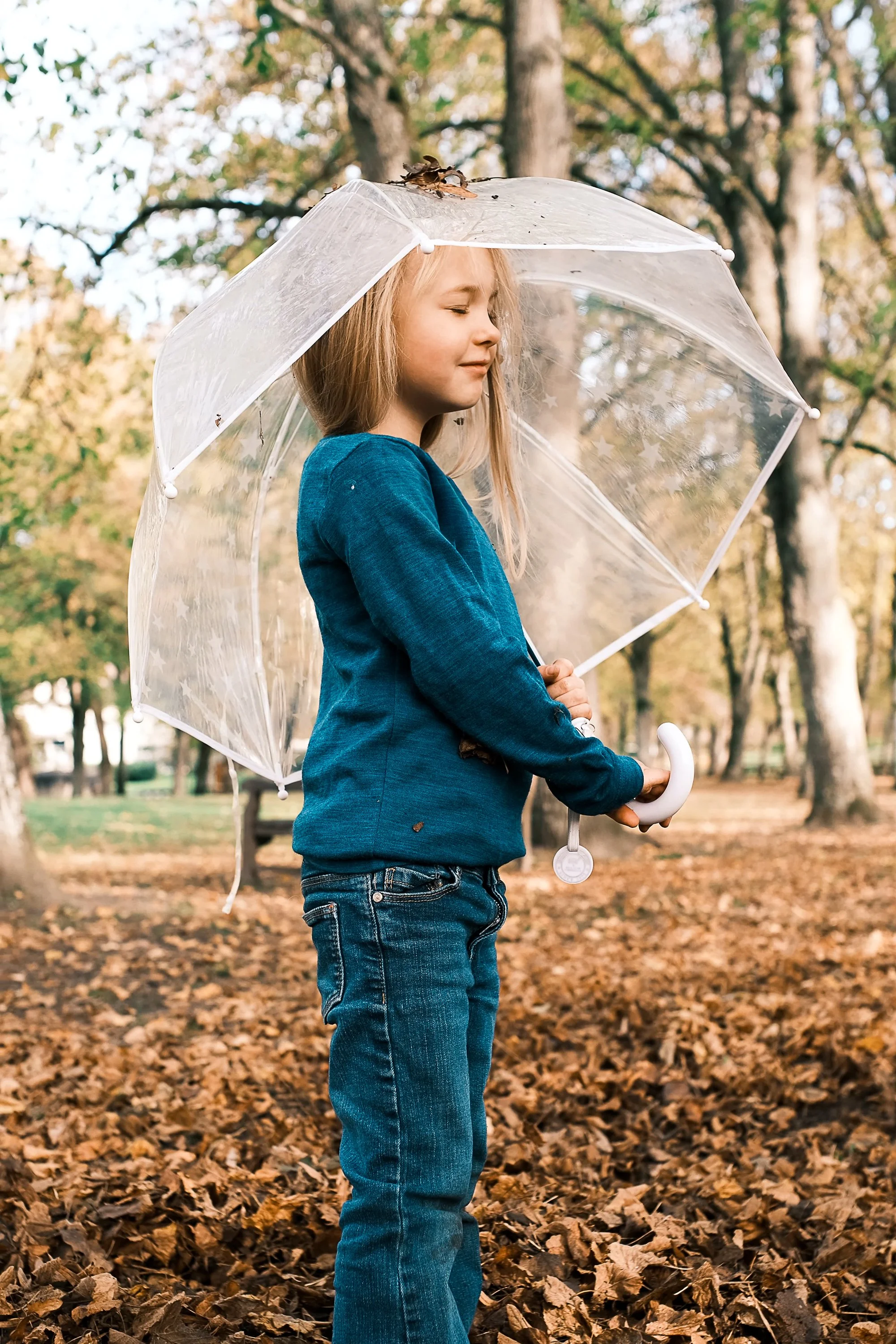 A young girl with blonde hair holding a transparent umbrella outside in an autumn park with fallen leaves. outdoor product photography children's fashion brand photographer childrenswear clothing