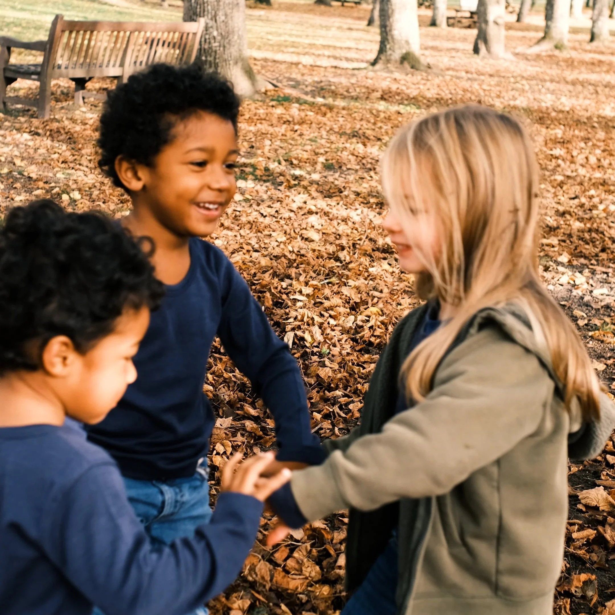 Three children playing and holding hands in a park with fallen autumn leaves and trees in the background. outdoor product photography children's fashion brand photographer childrenswear clothing