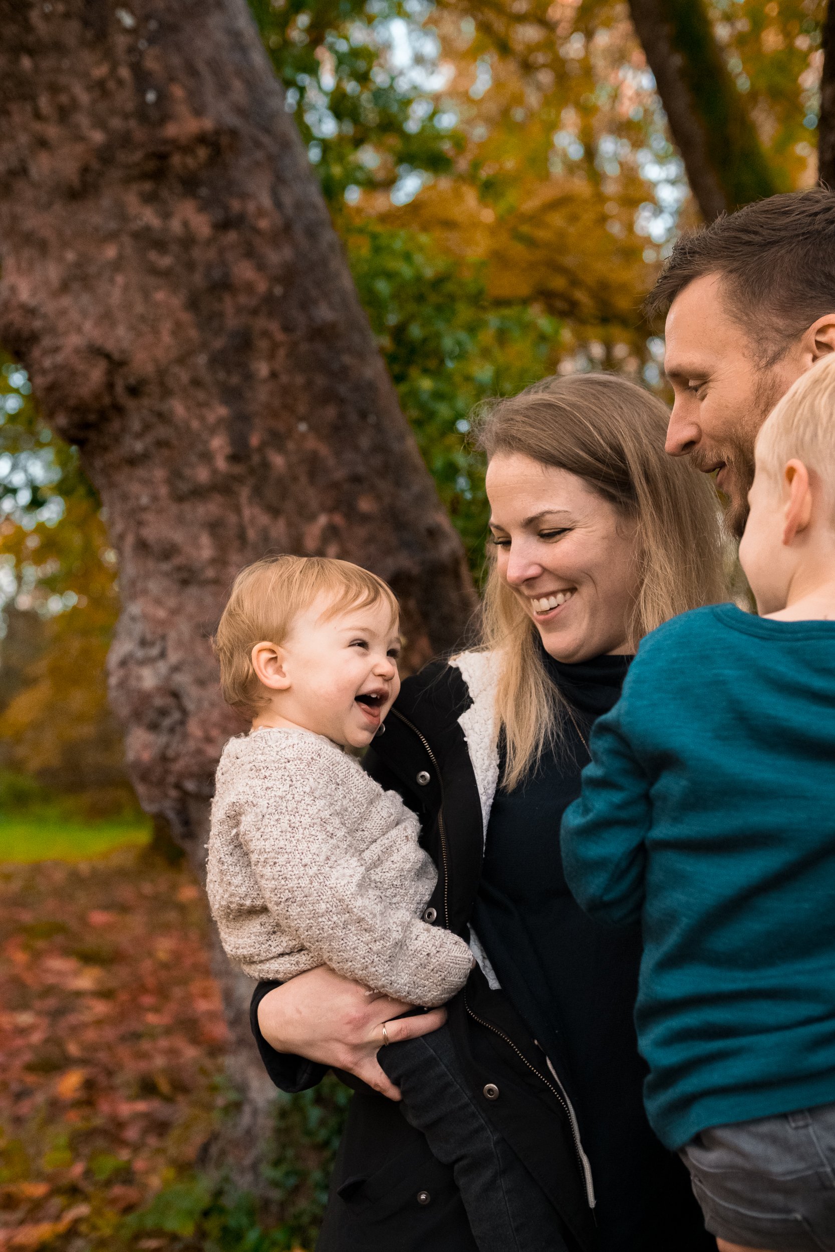 A family of four, including a woman, a man, and two young children, are smiling and playing outdoors in a park with autumn foliage. Product photography fashion photography for childrens brands.