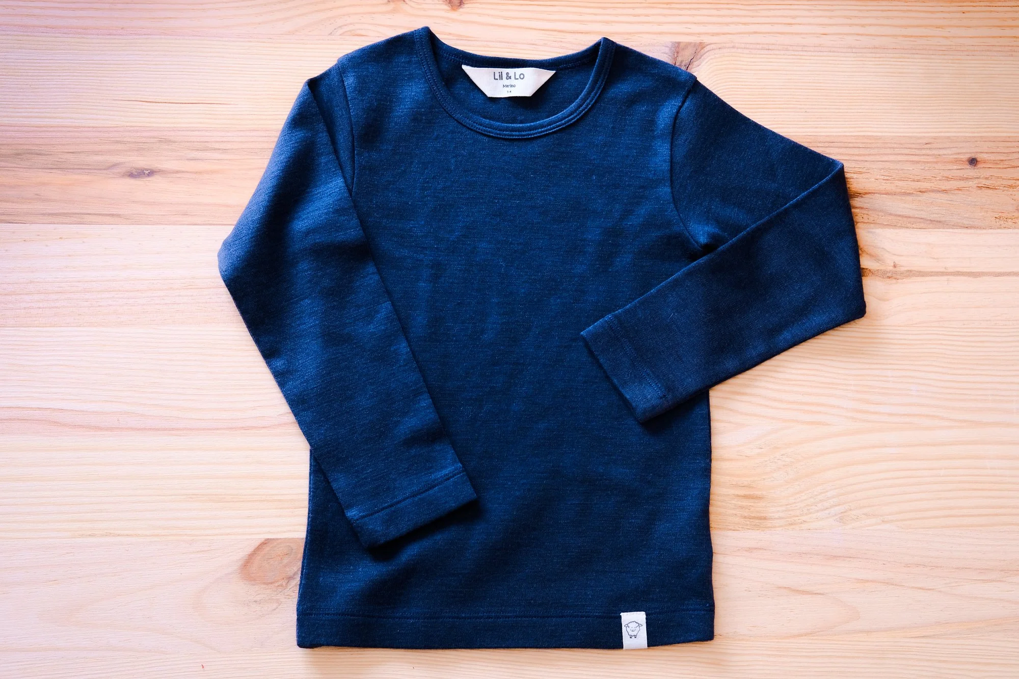 A blue long-sleeve shirt with a white brand label on the collar, laying flat on a light wooden surface. Product photography fashion photography for childrens brands.