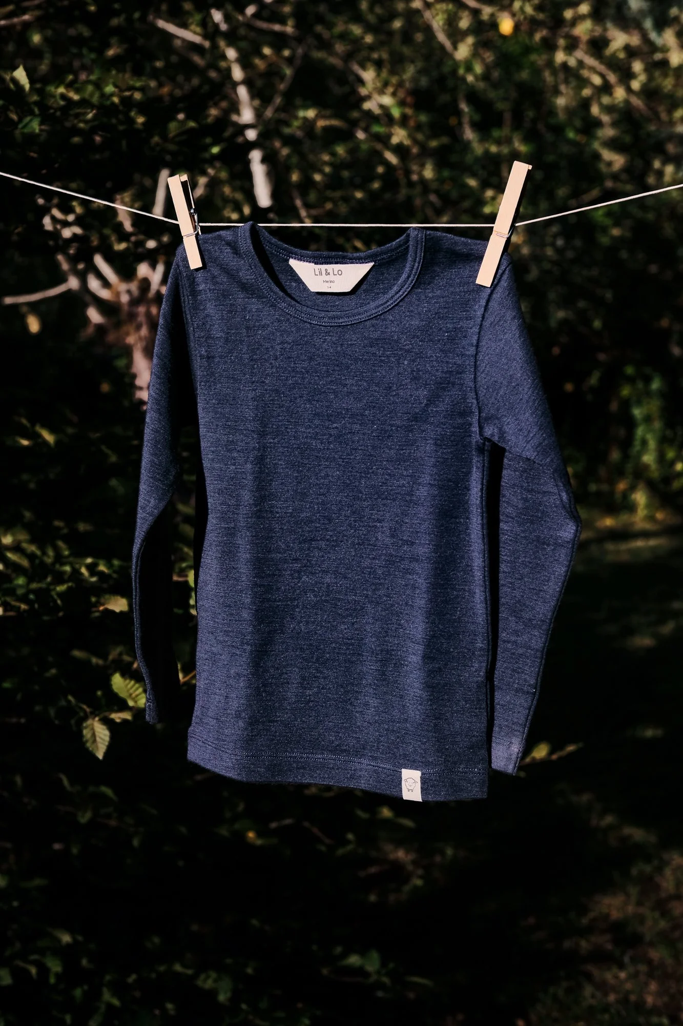 A dark blue long-sleeve shirt hanging on a clothesline with clothespins, outdoors with trees in the background. Fashion photography for childrens brands.