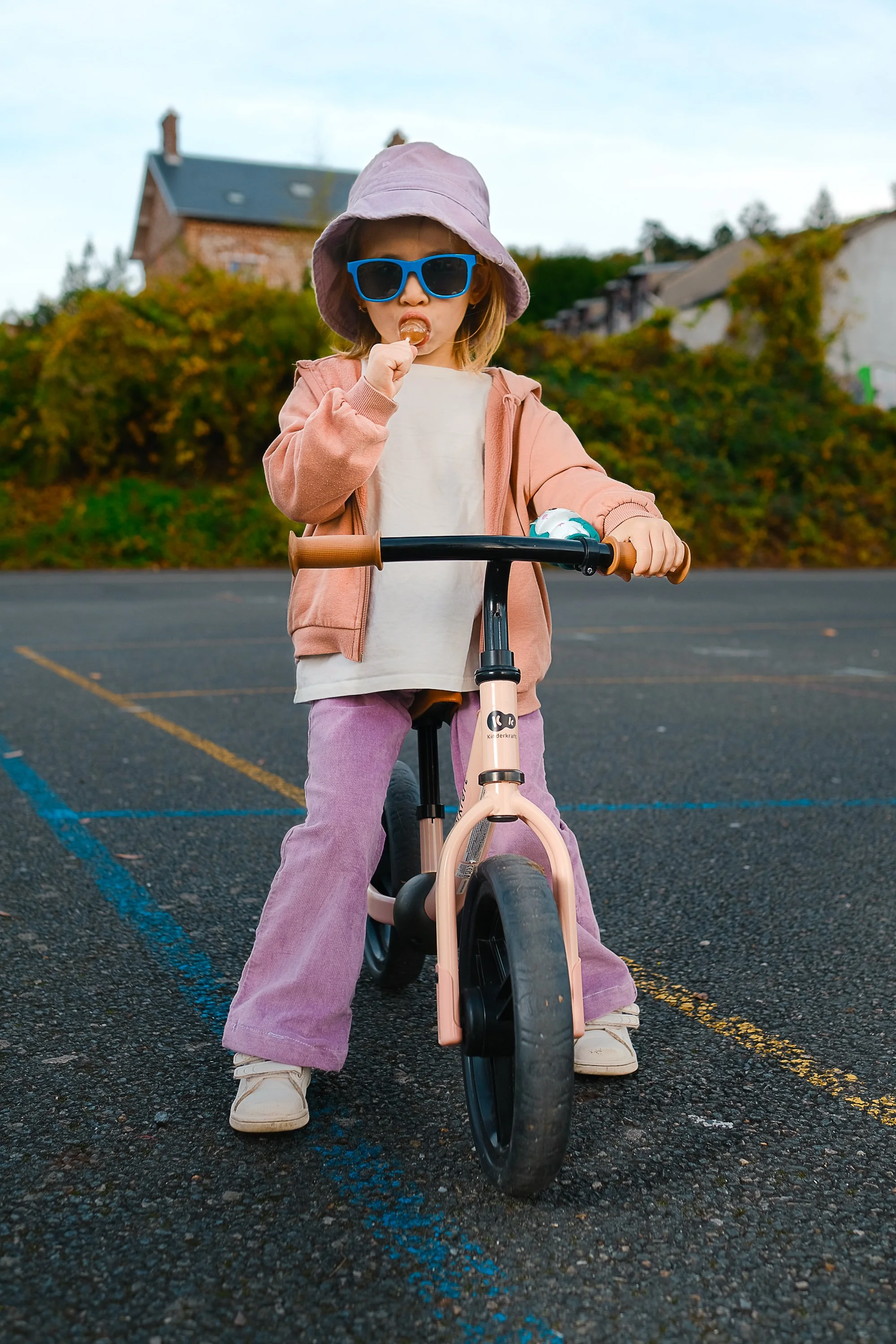 Young girl wearing sunglasses and a sun hat is riding a bike in an outdoor parking lot, eating a snack. Product photography fashion photography for childrens brands.