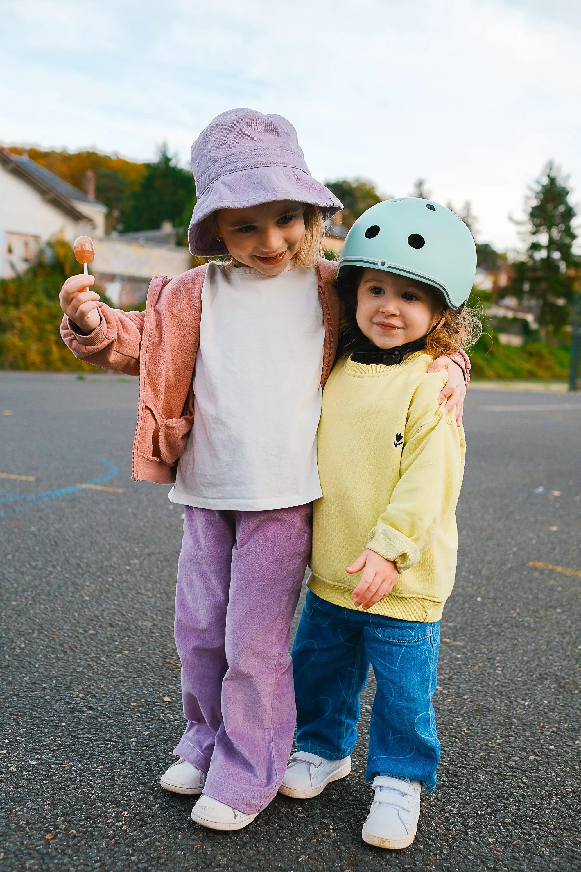 Two young girls hugging outdoors on a court, one with a purple hat holding a lollipop and the other wearing a bike helmet and yellow sweater. Editorial product photography fashion photography for childrens brands.
