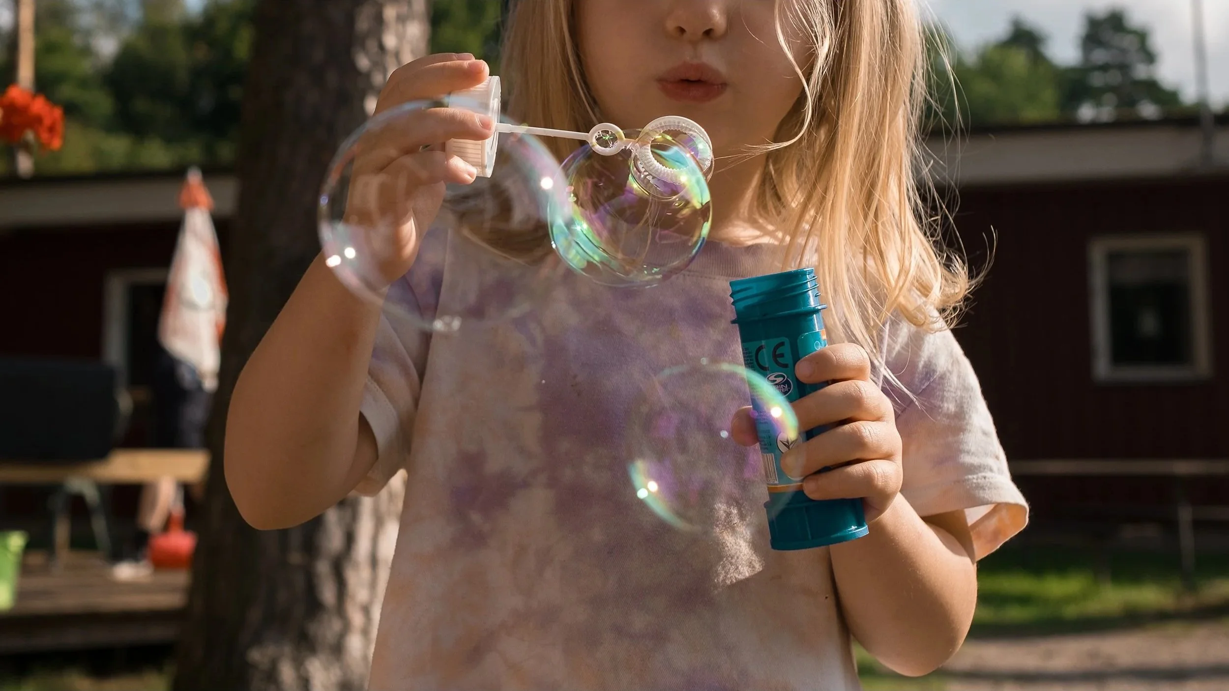 Child blowing soap bubbles outdoors, holding a plastic bottle of bubble solution, with a house and trees in the background. outdoor product photography children's fashion brand photographer childrenswear clothing