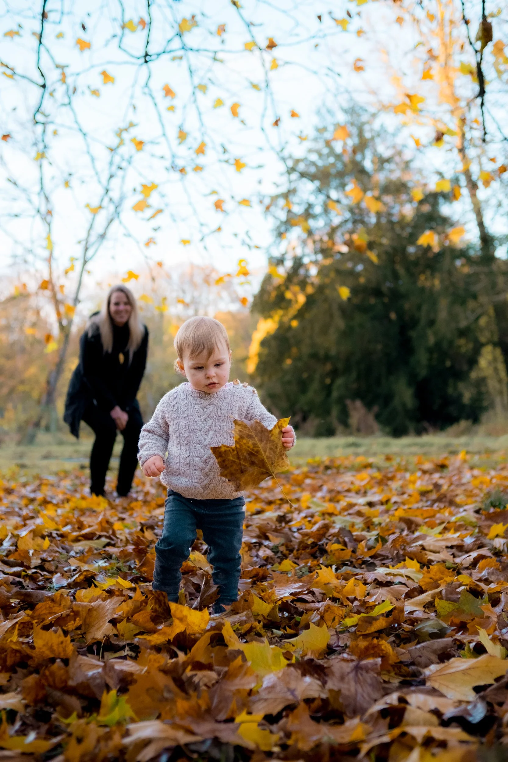 A young child wearing a beige knitted sweater and dark pants is playing among fallen autumn leaves, holding a large leaf in their hand, with a woman smiling and watching in the background. Product photography fashion photography for childrens brands.