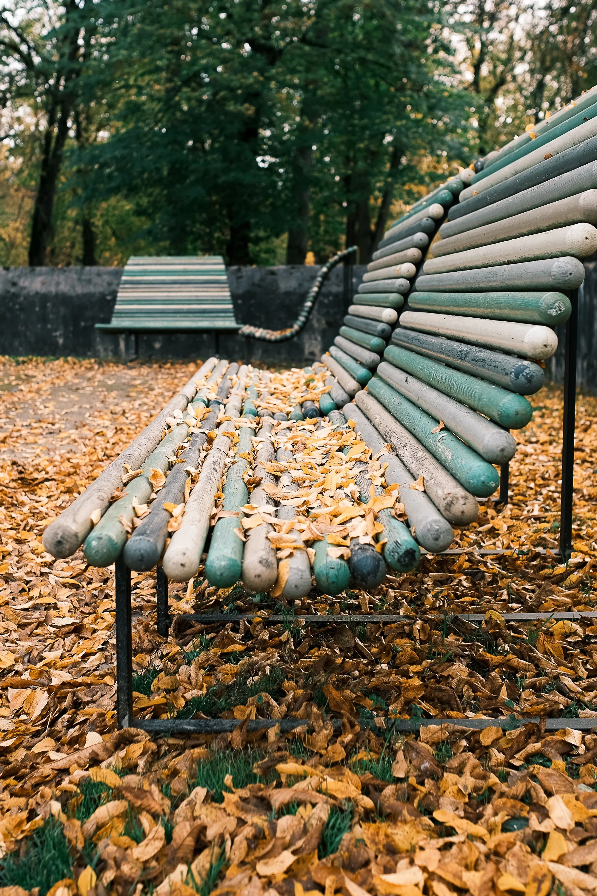 A colorful outdoor bench made of horizontal wooden logs with fallen autumn leaves on and around it, set in a park with trees in the background. outdoor product photography children's fashion brand photographer childrenswear clothing