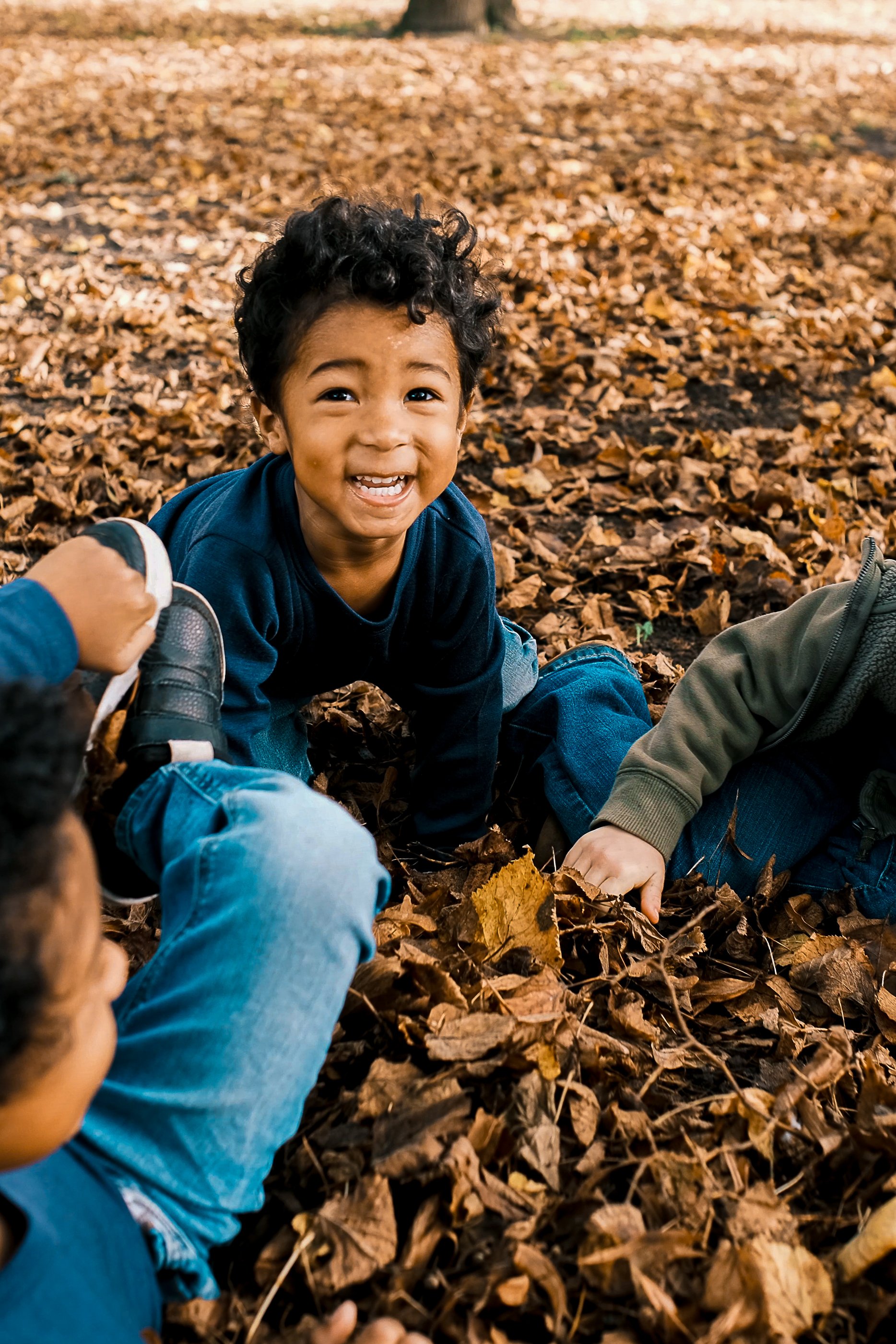 Young boy with curly hair smiling and playing in a pile of fallen autumn leaves, with other children partially visible around him. Product photography fashion photography for childrens brands.