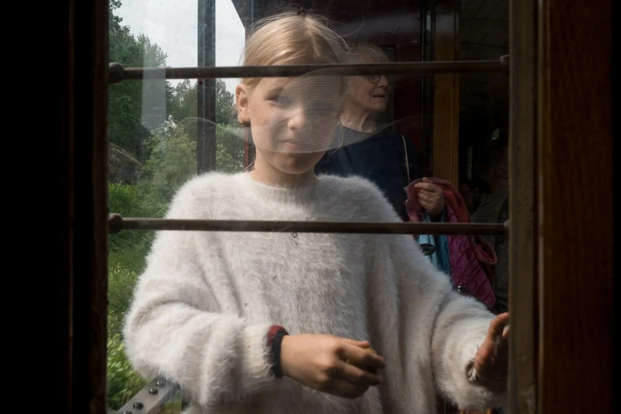 A girl with blonde hair and a white fuzzy sweater is looking out through a window with horizontal bars, with a reflection of her face on the glass. streetwear children's fashion brand photographer