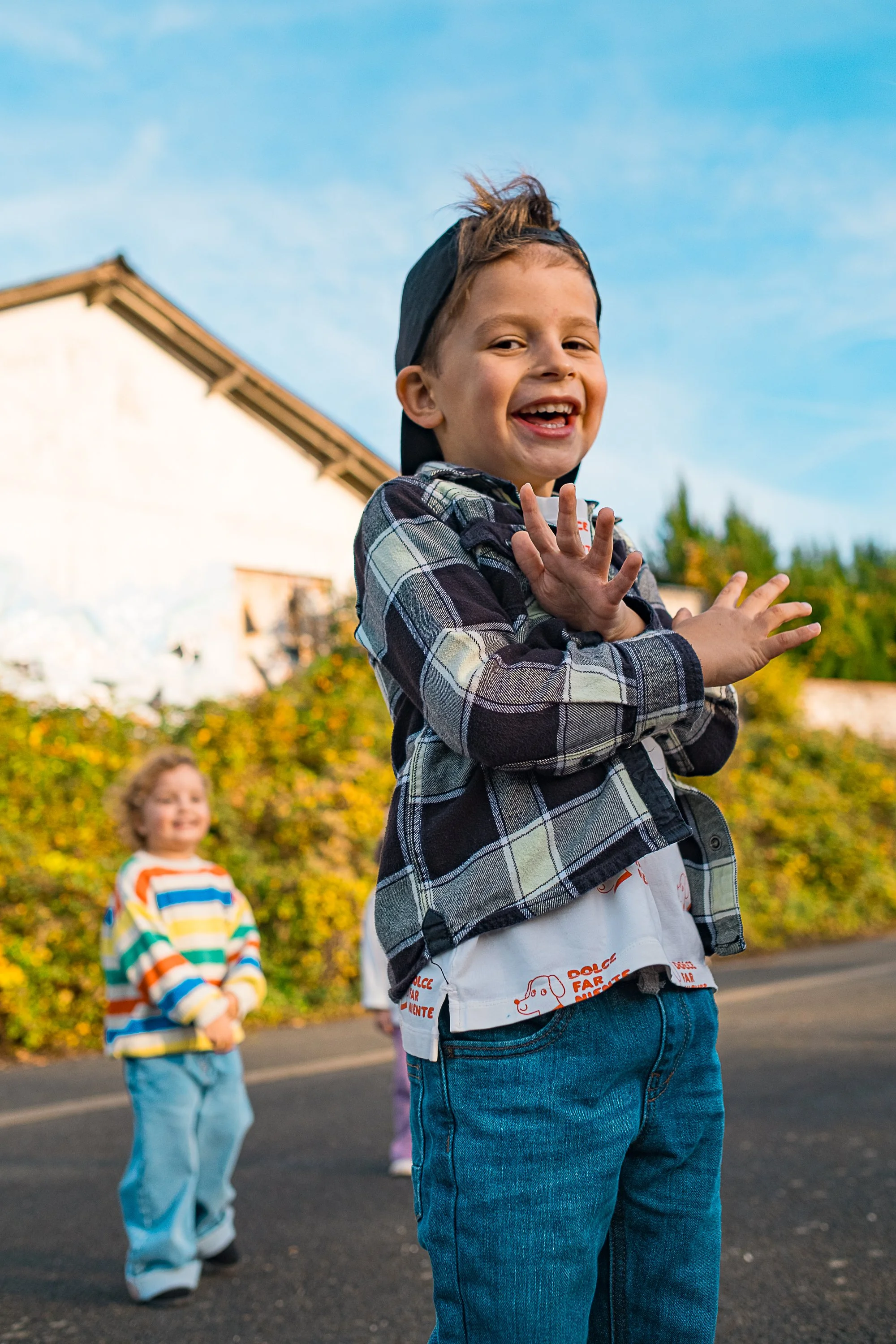 A young boy in a baseball cap and checkered jacket smiling and waving at the camera, with two children in the background outdoors on a sunny day. Product photography fashion photography for childrens brands.