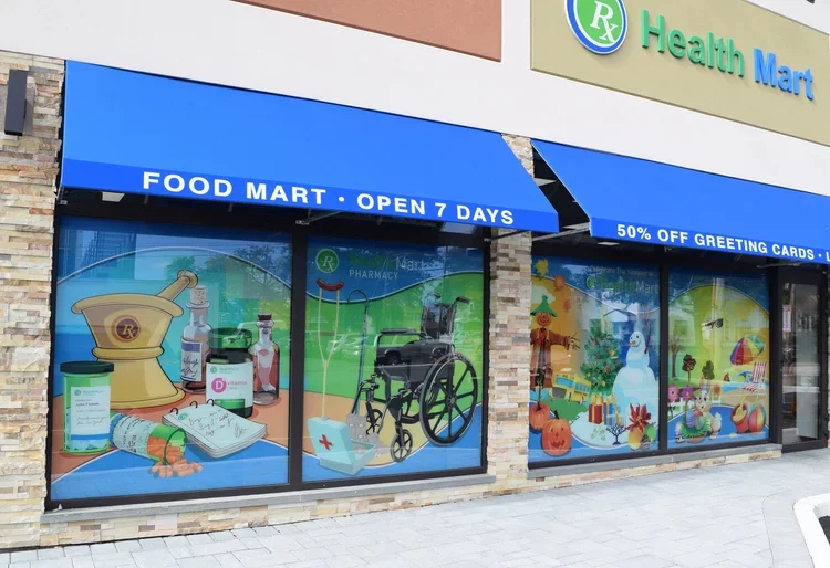 Exterior storefront of a grocery store called Food Mart, open 7 days a week. The store has large windows with colorful illustrations of health and wellness items, including medications, a wheelchair, and holiday-themed decorations. Blue awning with w