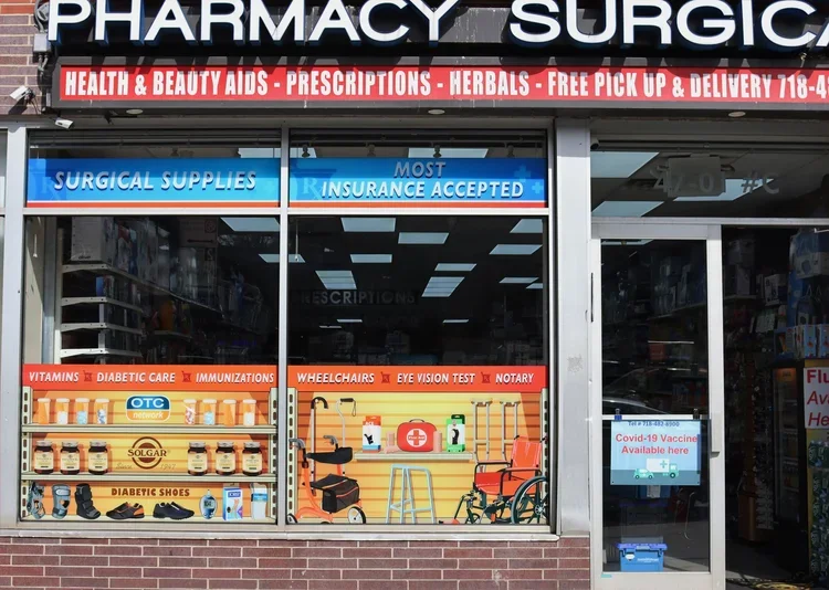 View of a pharmacy storefront with signs advertising health and beauty aids, prescriptions, herbals, free pickup and delivery, and accepting most insurance. The window displays medical supplies, diabetic shoes, vitamins, diabetic care, immunizations,