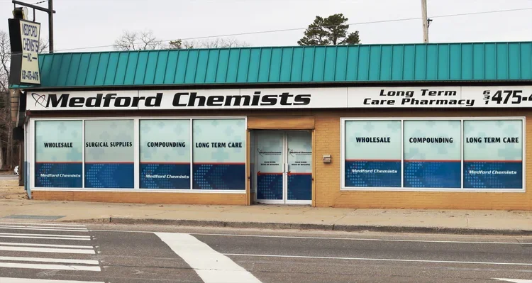 Storefront of Medford Chemists with signs for wholesale, surgical supplies, compounding, and long-term care pharmacy, located on a street with crosswalk markings.