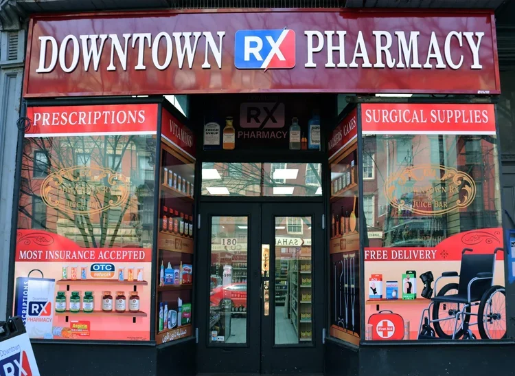 Facade of a pharmacy storefront labeled 'Downtown RX Pharmacy' with signs indicating prescriptions, surgical supplies, and free delivery.