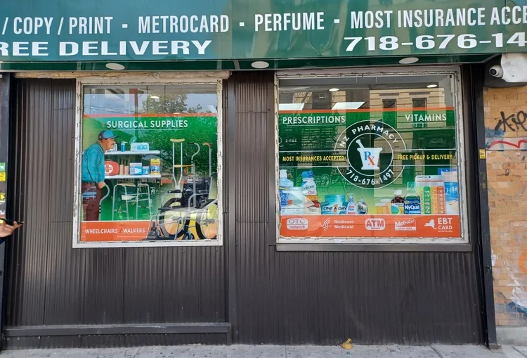 Front of a local pharmacy and drugstore with signs for prescription medications, vitamins, surgical supplies, and insurance services. The window displays various health products, and there is a person standing inside near shelves.