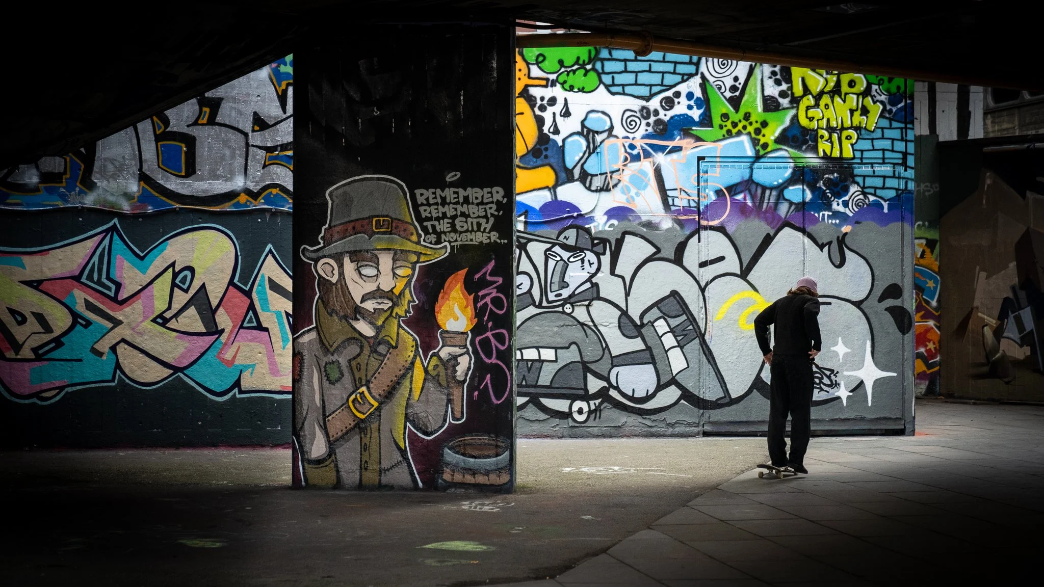 A skateboarder near colorful graffiti murals on a city wall, featuring characters and abstract art under a bridge or overpass.