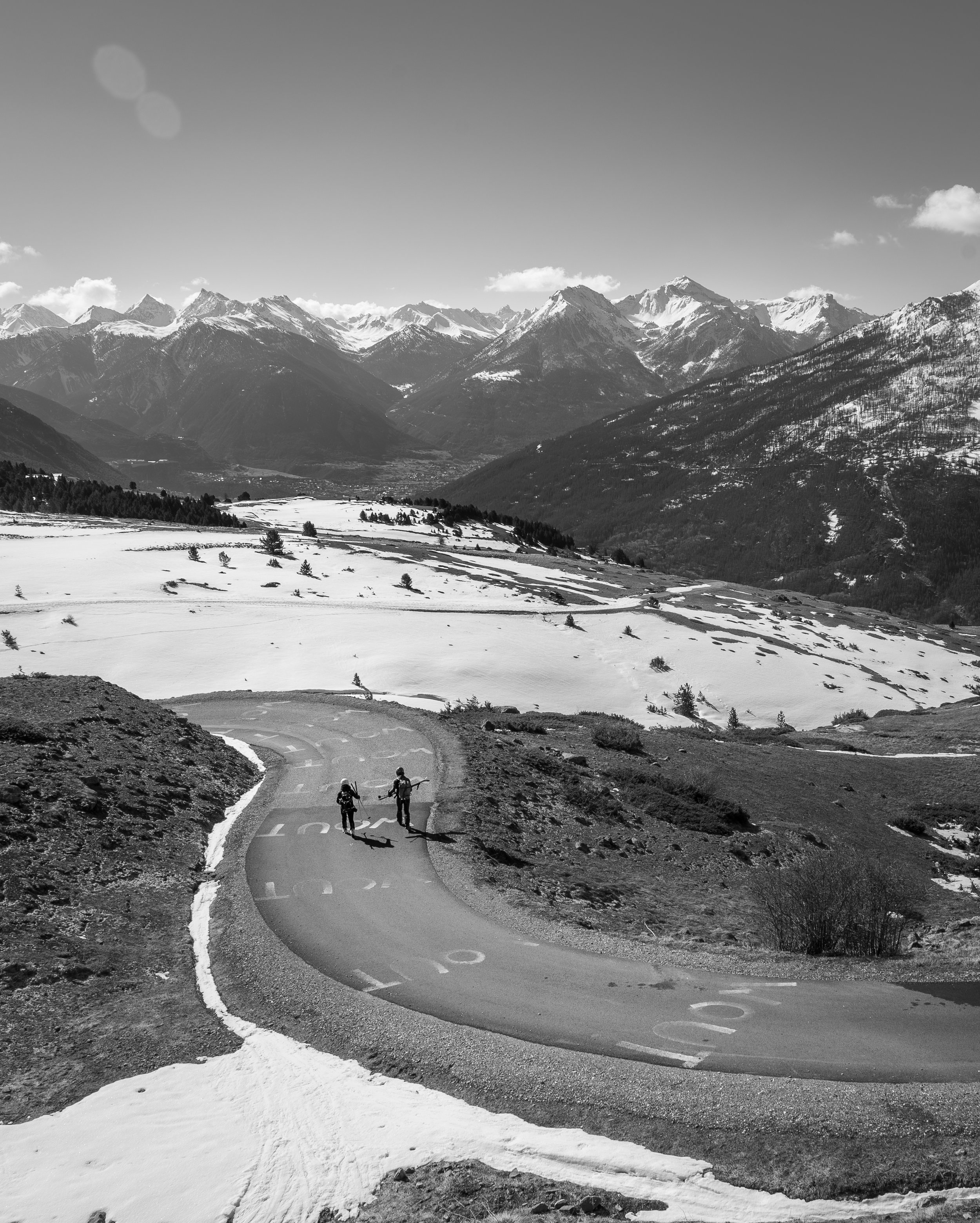Two hikers walking on a winding mountain road with snow-covered landscape and mountain peaks in the background
