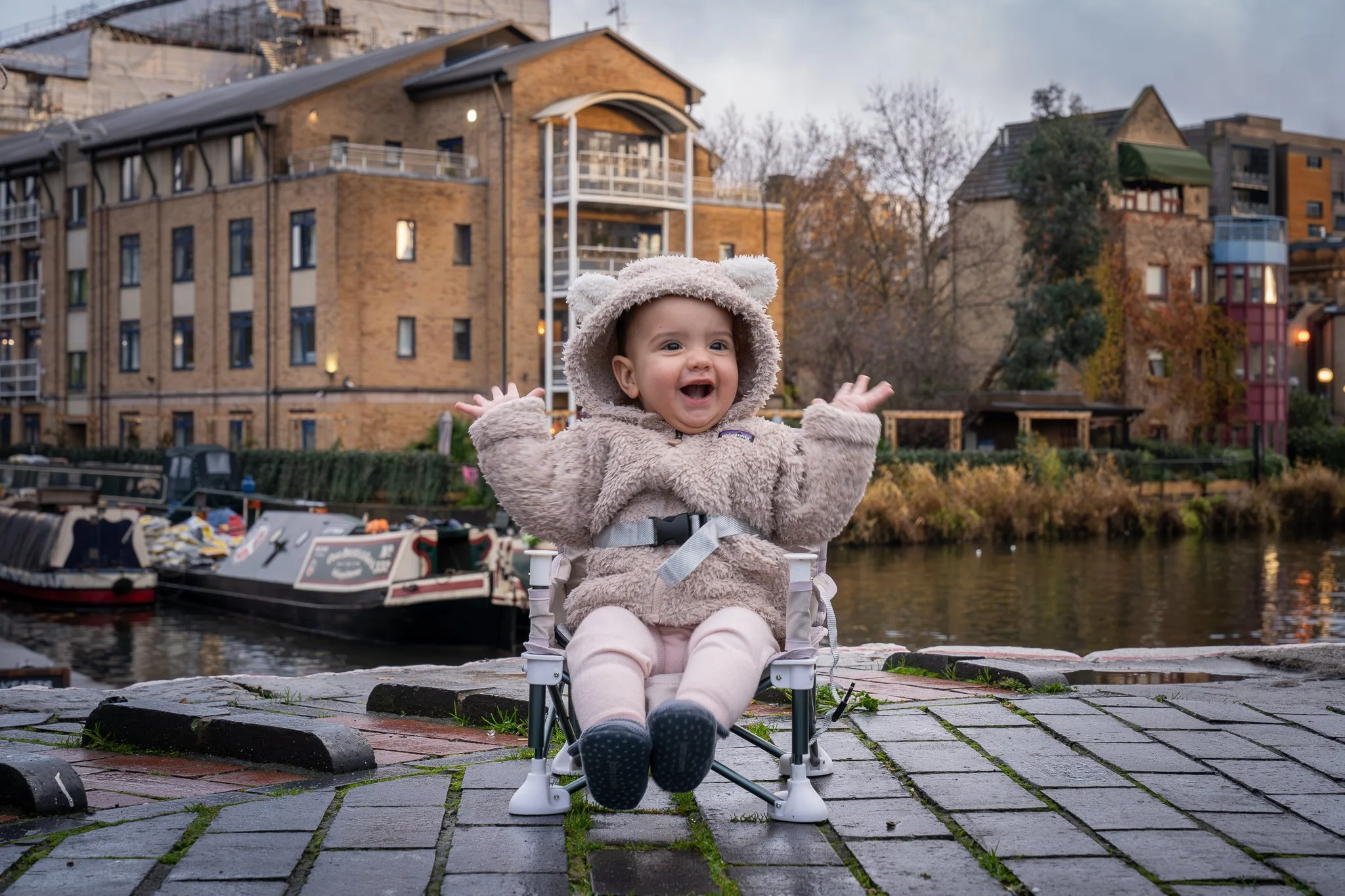 A baby dressed in a cozy bear costume with a hood featuring bear ears, sitting in a small chair by a canal, smiling and waving. The background shows boat houses and apartment buildings with autumn trees.