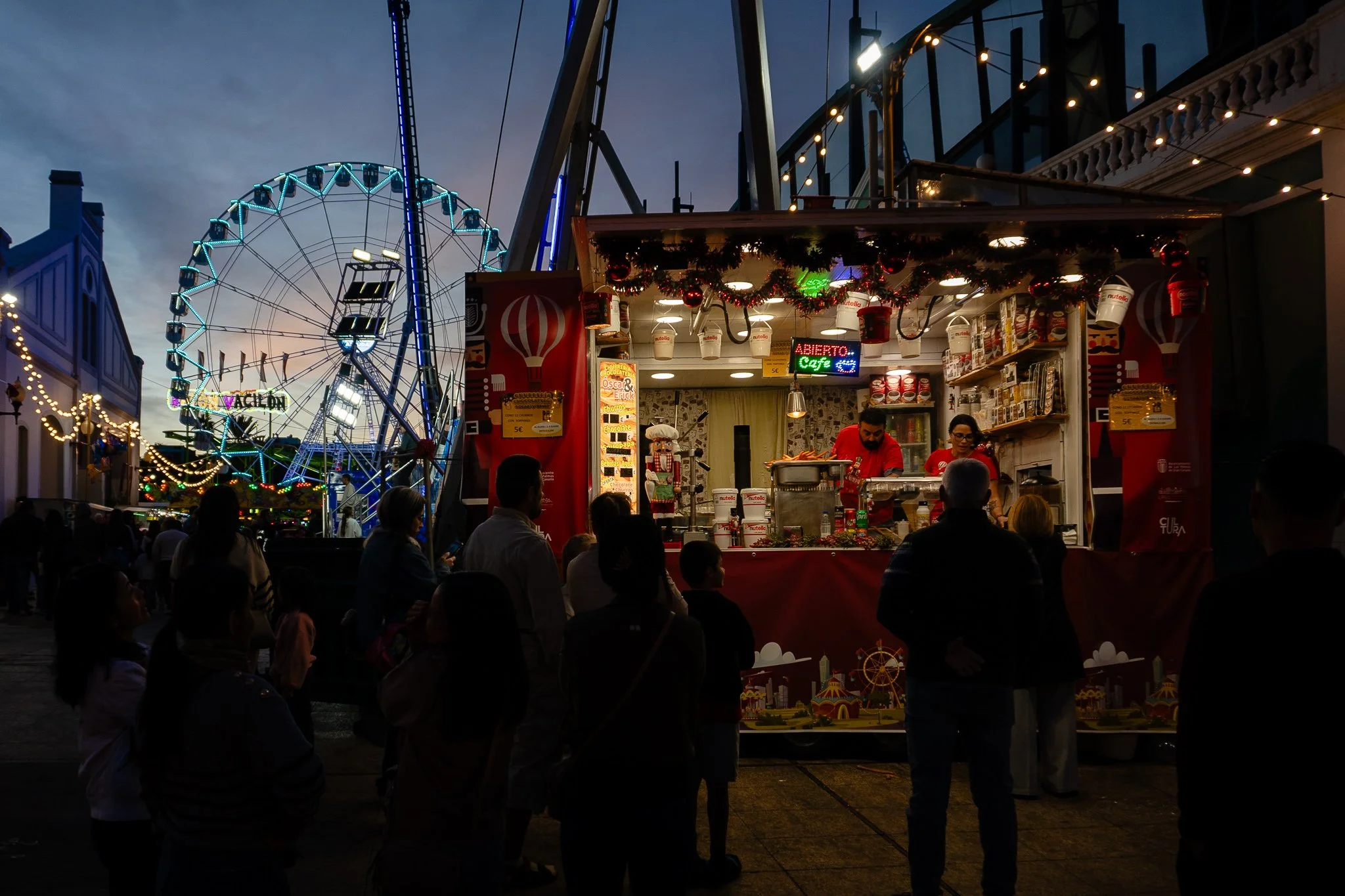 Nighttime scene at a fair with a large Ferris wheel in the background, colorful lights, and a small food stand in the foreground serving popcorn or similar snacks, with people gathered around.