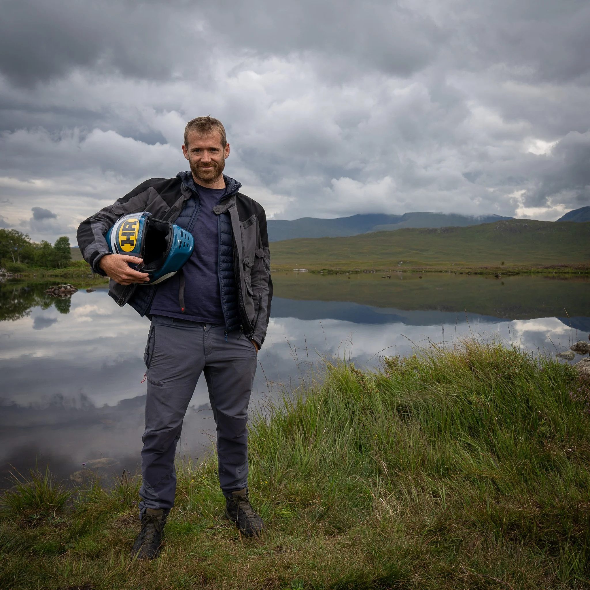A man standing outdoors near a lake with mountains and cloudy sky in the background, holding a motorcycle helmet.