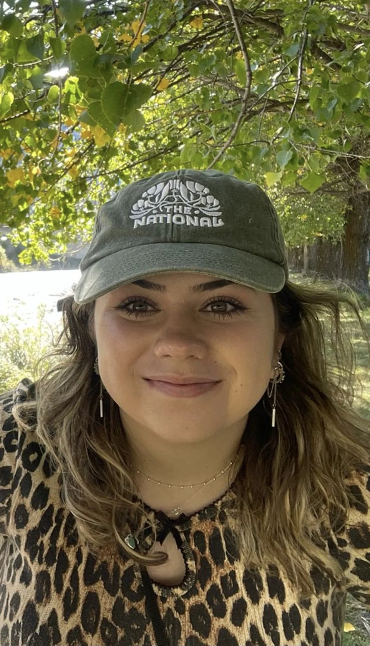 A woman smiling outdoors, wearing a gray cap with 'The National' logo, earrings, a delicate necklace, and a leopard print top, with green trees and sunlight in the background.