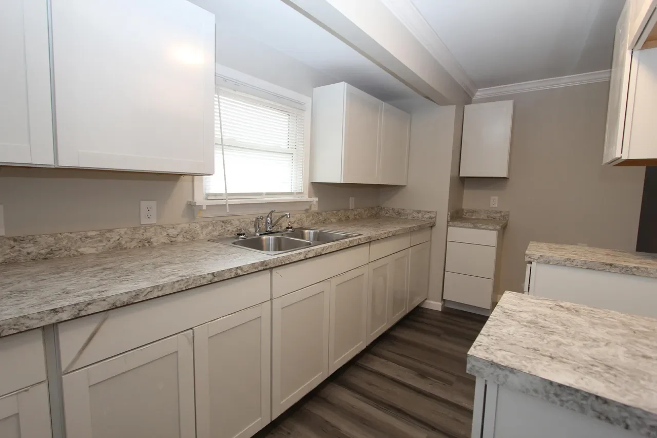Kitchen with white cabinets, granite countertops, a double sink, and a window above the sink. There is wood-like flooring and an empty countertop on the right.