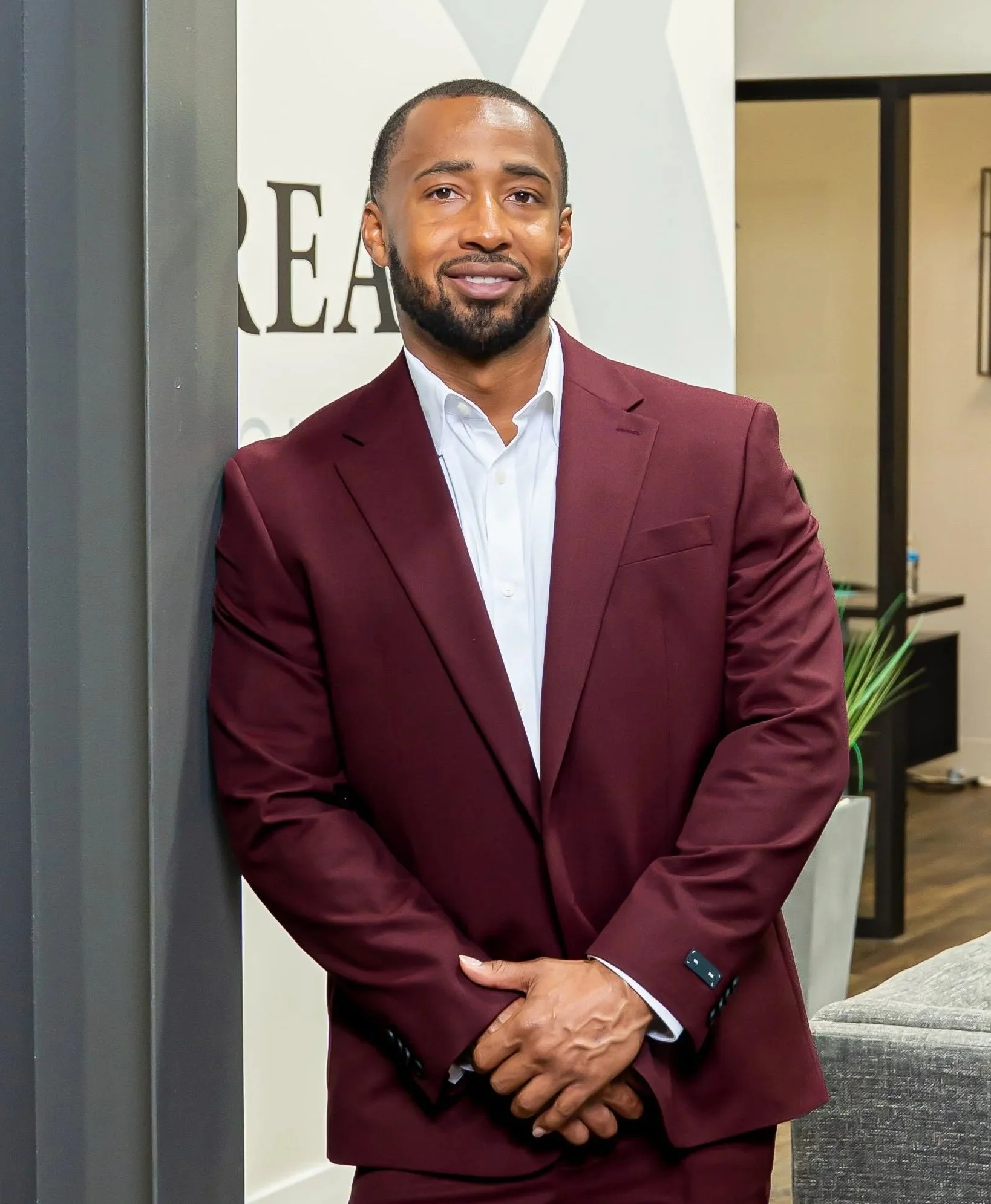 Lance Morton wearing a white shirt and maroon suit jacket, standing indoors and leaning against a wall.