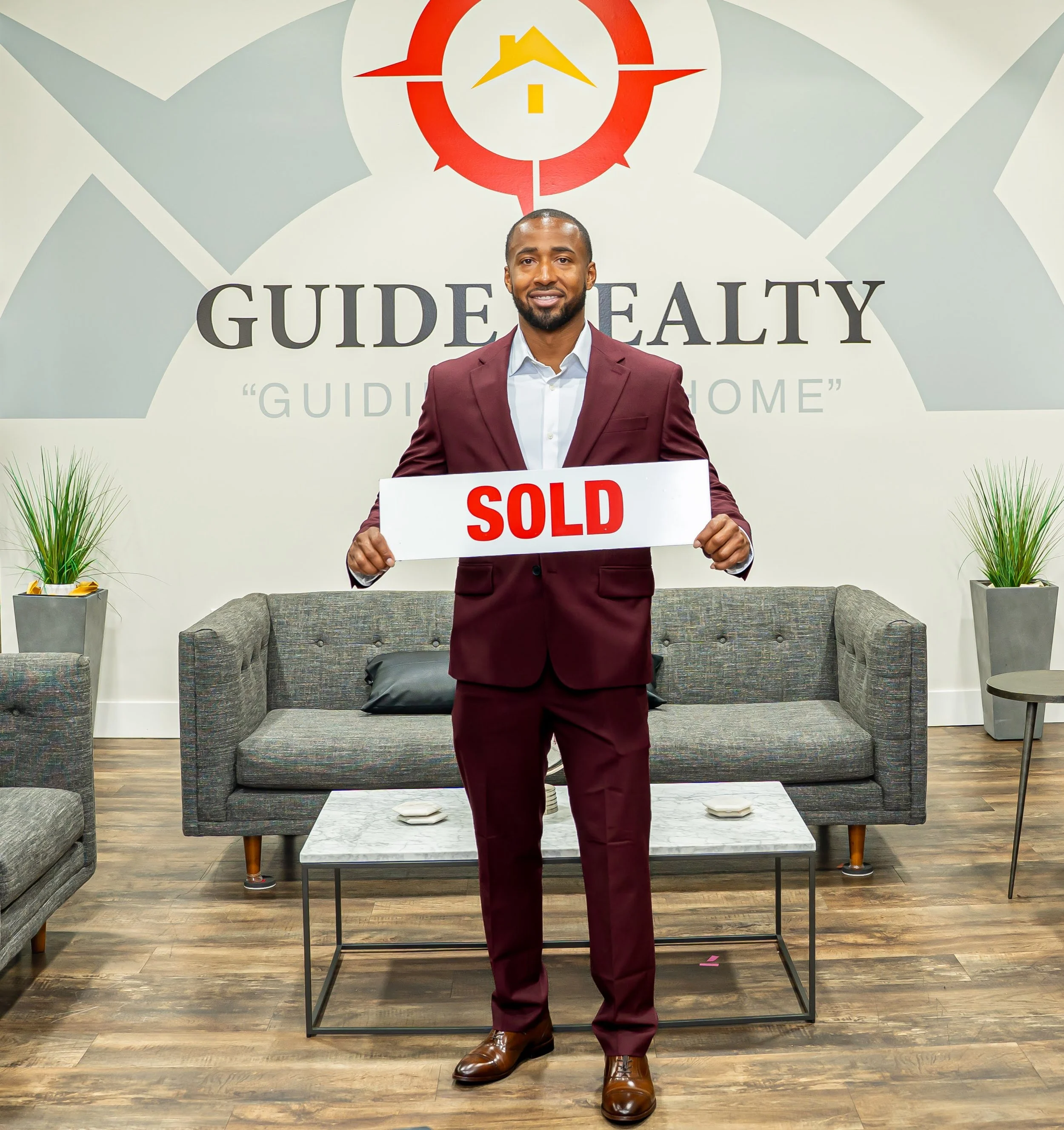 Lance Morton wearing a maroon suit while holding a 'SOLD' sign inside a real estate office, with a wall display behind him featuring a logo and the text 'GUIDE REALTY'.