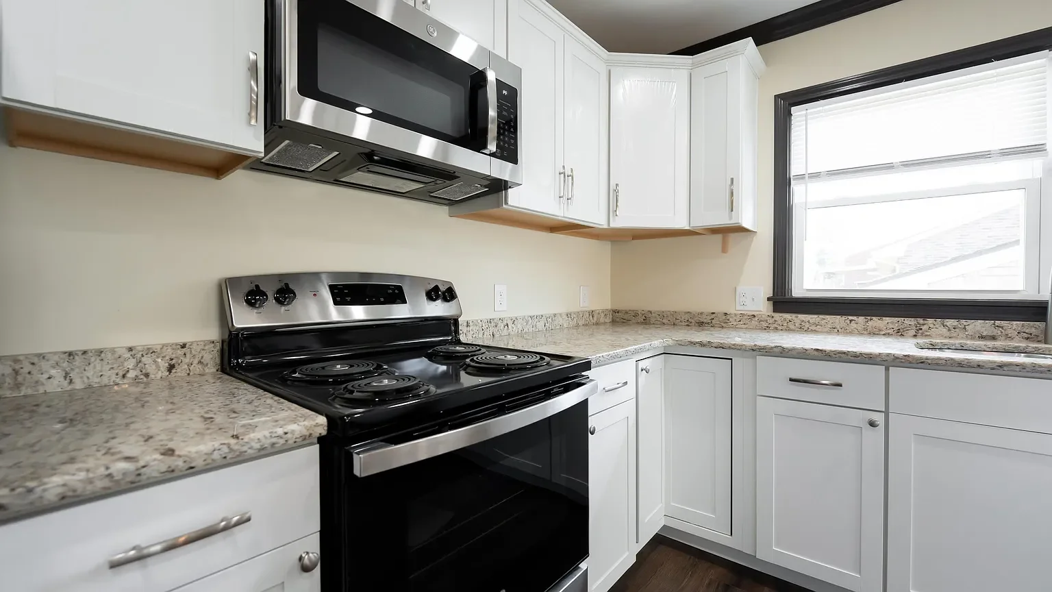 Kitchen with white cabinets, a granite countertop, a black electric stove, a microwave above, a window with blinds, and a wooden shelf.