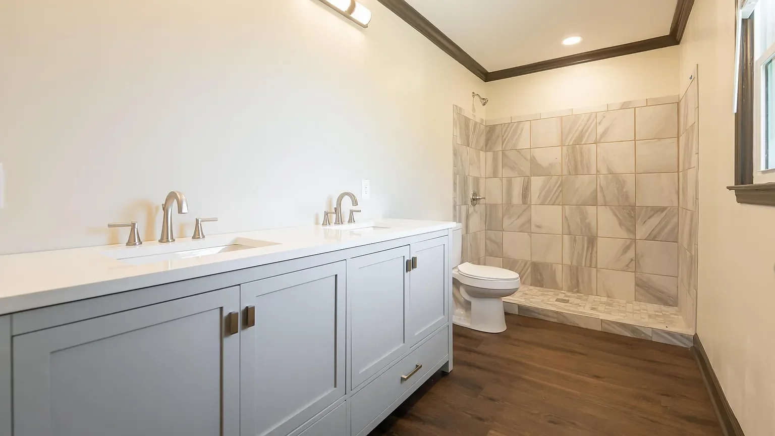 A clean, modern bathroom with a white double vanity, silver faucets, a toilet, and a walk-in shower with beige tiles and a small window.