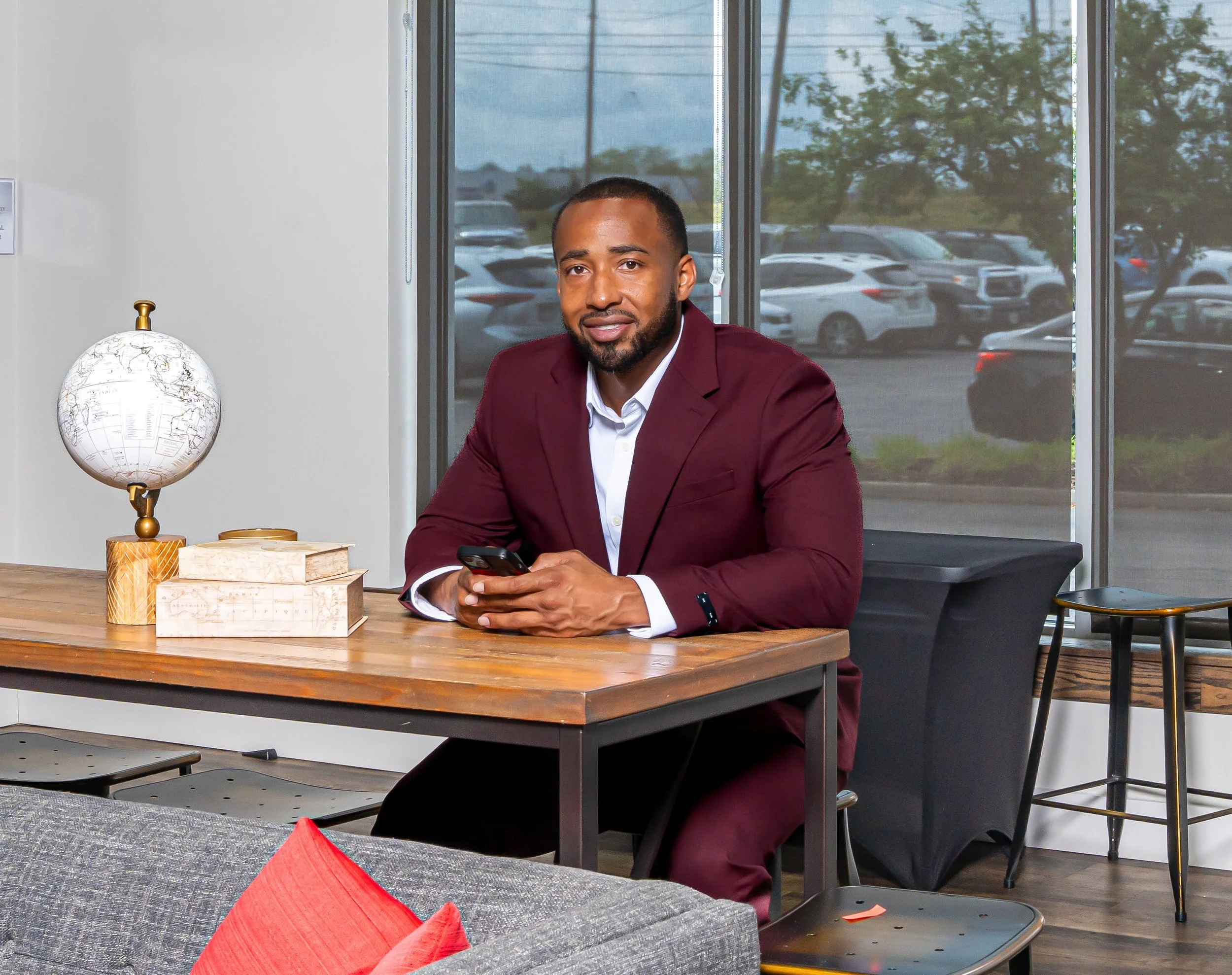 Lance Morton wearing a burgundy suit and white shirt sits at a wooden desk inside an office, holding a smartphone. There is a globe and some books on the desk. Large windows show a parking lot and trees outside.