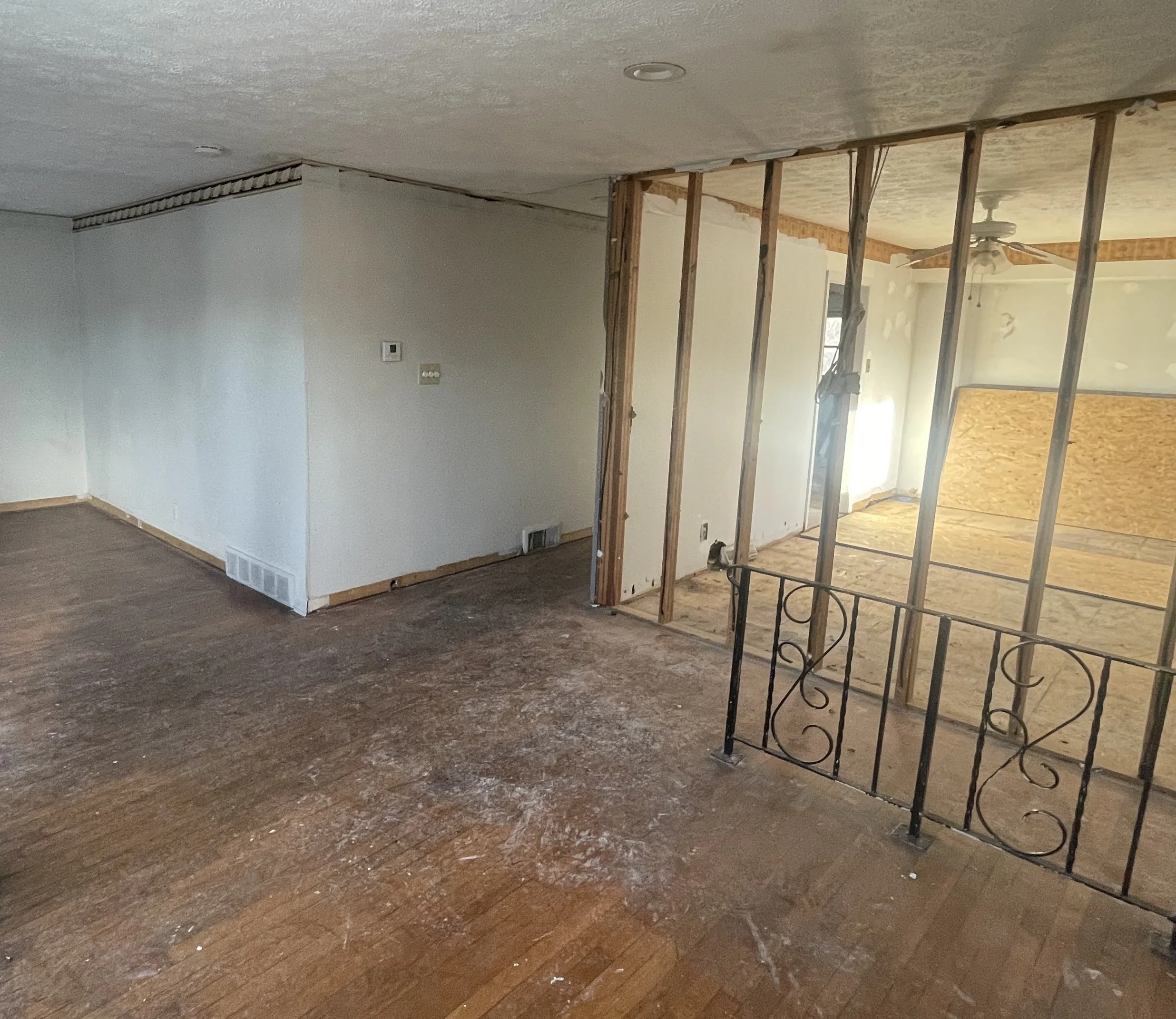 A room under renovation with partially removed wall, exposed wooden framing, and a dark, worn wooden floor.
