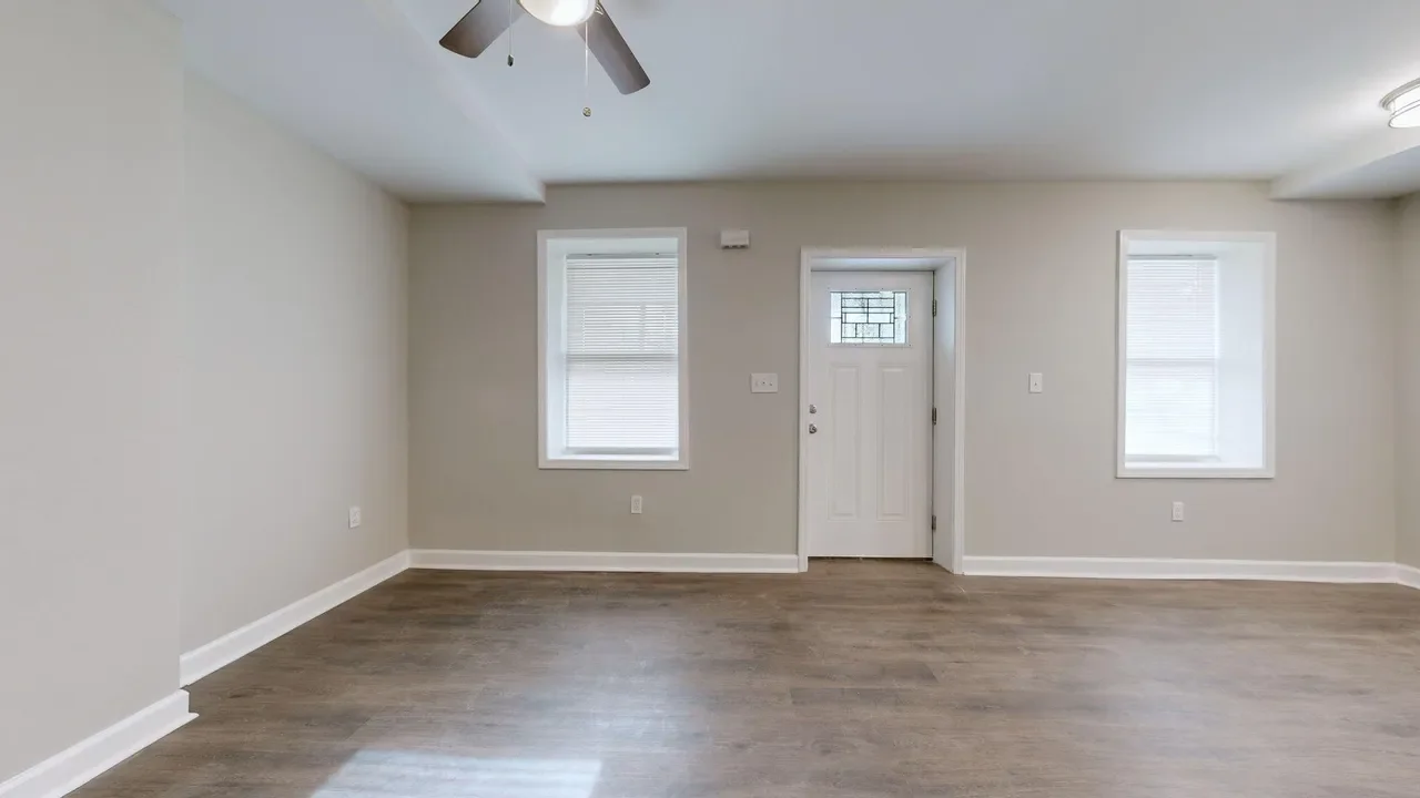 Empty living room with two windows, a front door, ceiling fan, and hardwood floors.