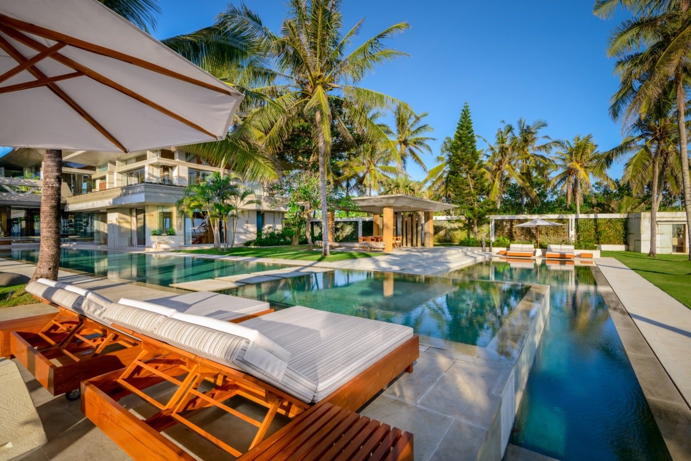 Luxury outdoor swimming pool area at a tropical resort with lounge chairs, umbrellas, palm trees, and a modern building in the background under a clear blue sky.