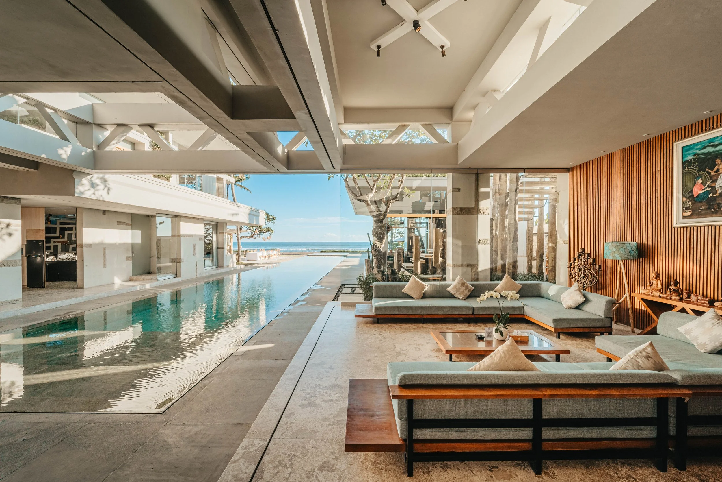 Modern lounge area with large glass windows overlooking a pool and ocean, featuring light gray sectional sofas with beige pillows, a wooden coffee table with a potted orchid, decorative items on a wooden sideboard, and artwork on a wooden paneled wal