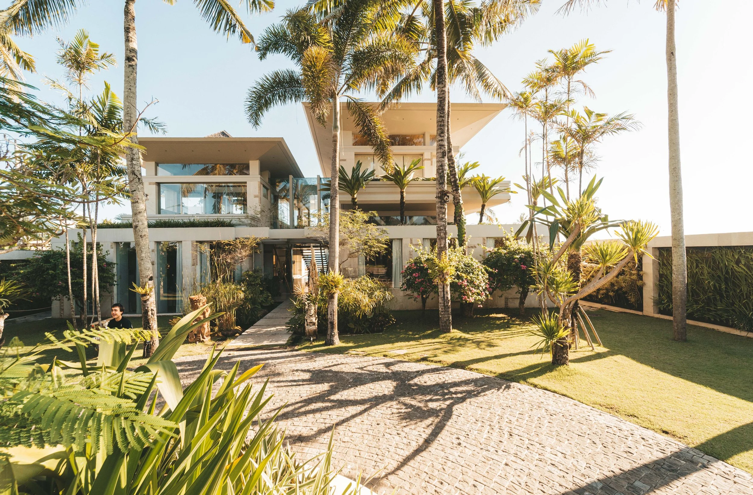 Modern house surrounded by palm trees and lush landscaping, with a stone pathway on a sunny day.