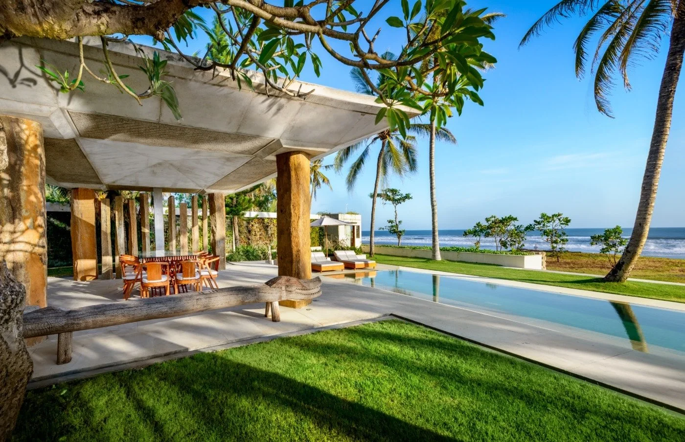 Luxury beachfront patio with outdoor dining table and chairs, lounge chairs with umbrellas, a swimming pool, palm trees, and ocean waves under a clear blue sky.