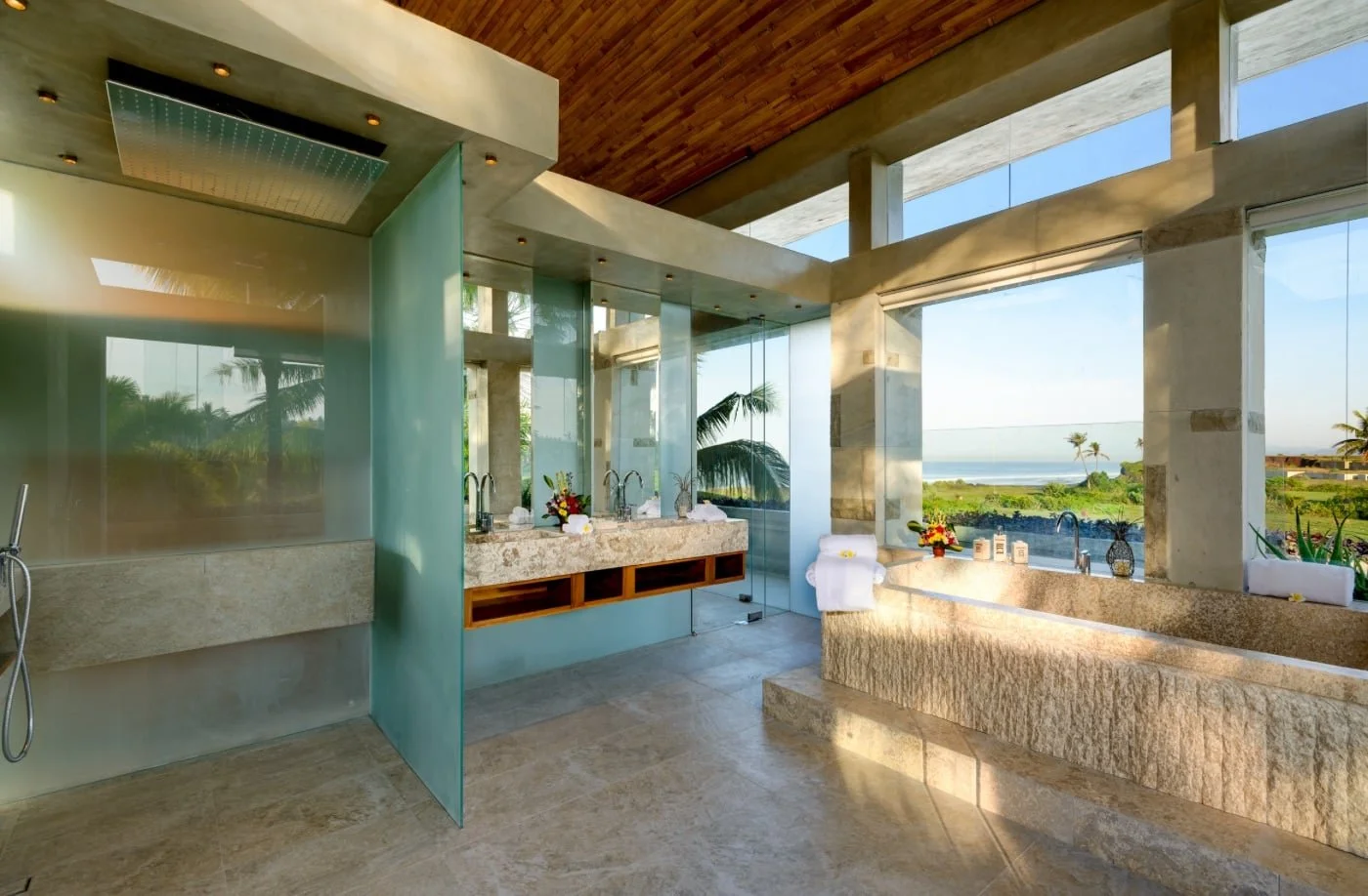 Luxurious bathroom with large windows showcasing a view of palm trees and the ocean, featuring a stone bathtub, double vanity with sinks, and a modern shower with a glass partition.
